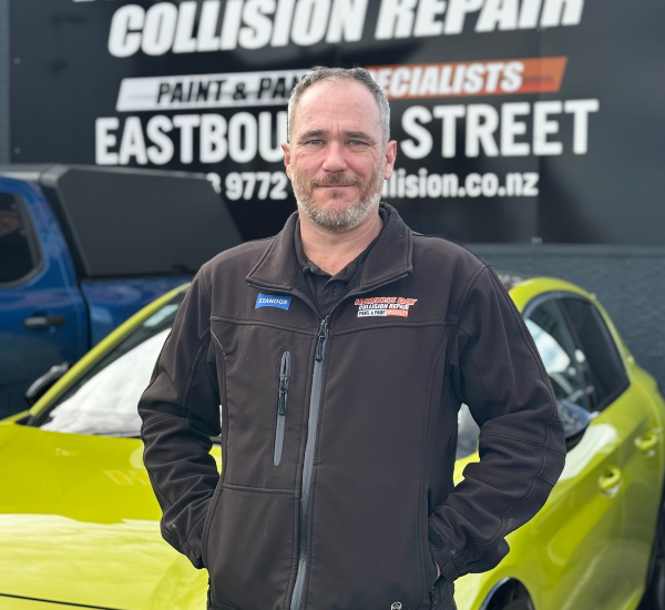 Man in a black jacket standing in front of a yellow sports car with a sign for an auto repair shop in the background.