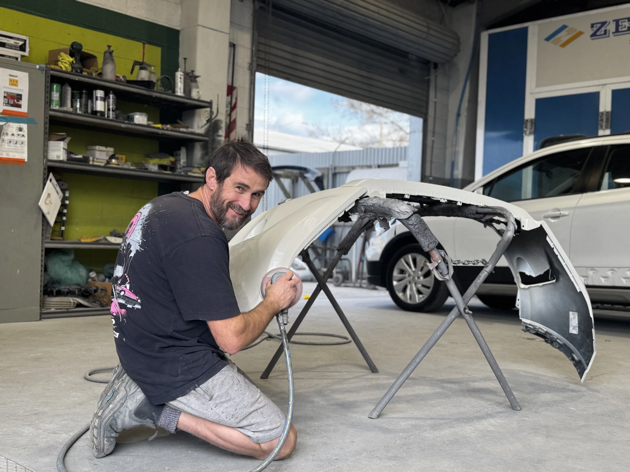 A man with a beard and dark hair, wearing a black T-shirt and gray shorts, kneeling on the floor and working on the frame of a car body in an auto repair shop.