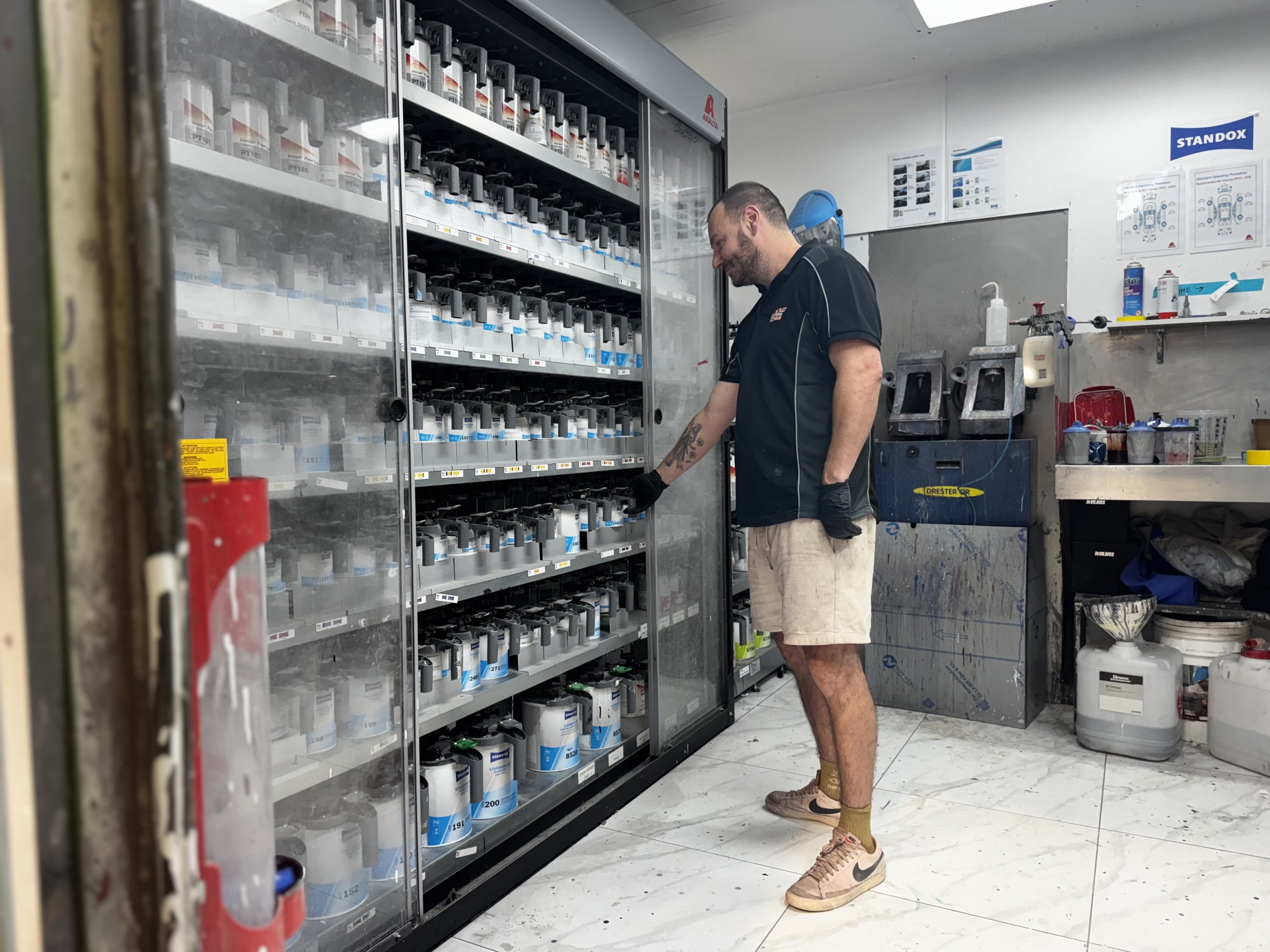 A man with a beard, wearing a black shirt, beige shorts, yellow socks, and tan sneakers, is looking at paint cans in a hardware store aisle. The aisle has a glass-fronted fridge with paint supplies and a workspace with various tools and posters on the wall.