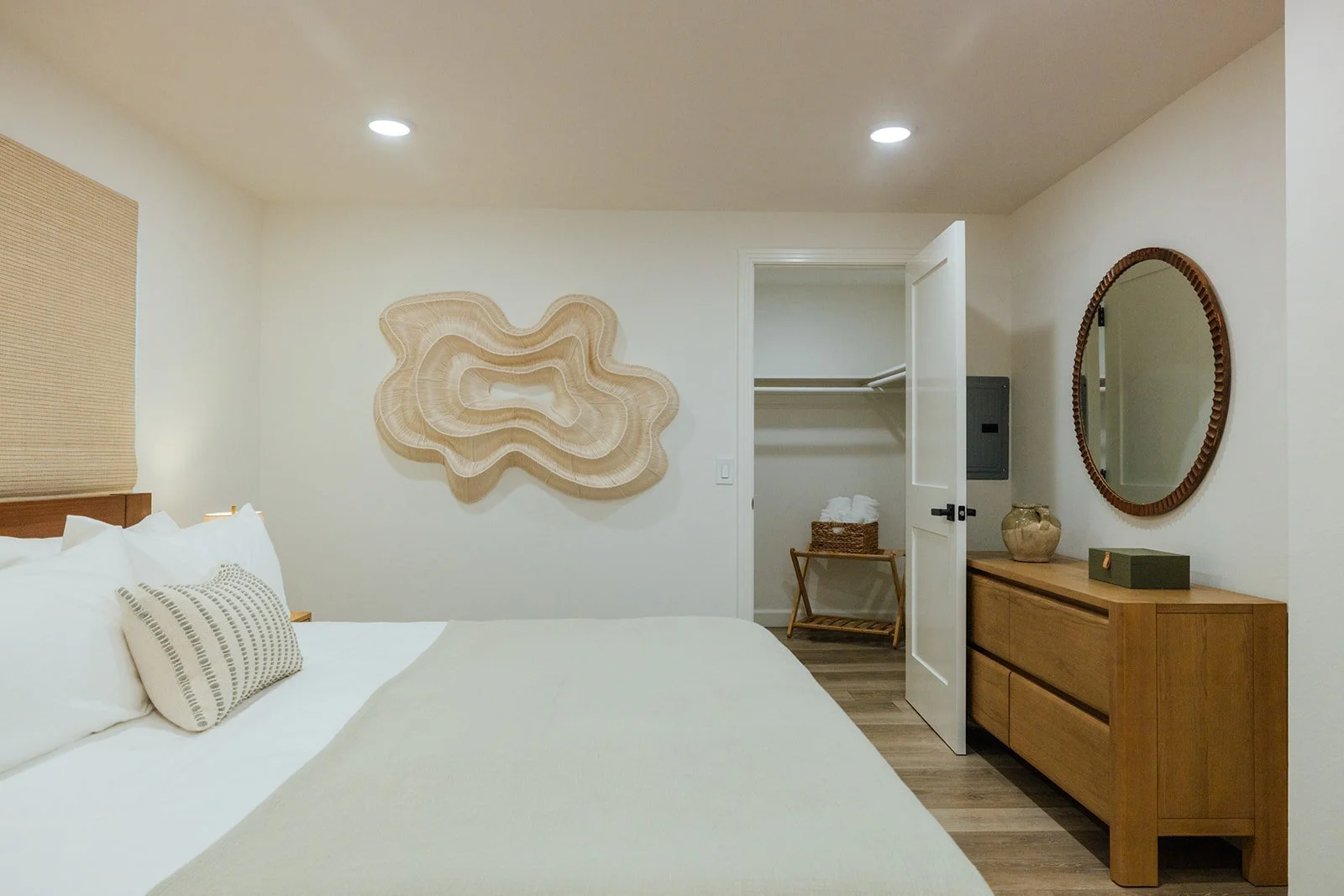 A cozy bedroom with a bed, beige pillows, a wall art piece, a mirror, a wooden dresser, an open closet with towels, and decorative items.