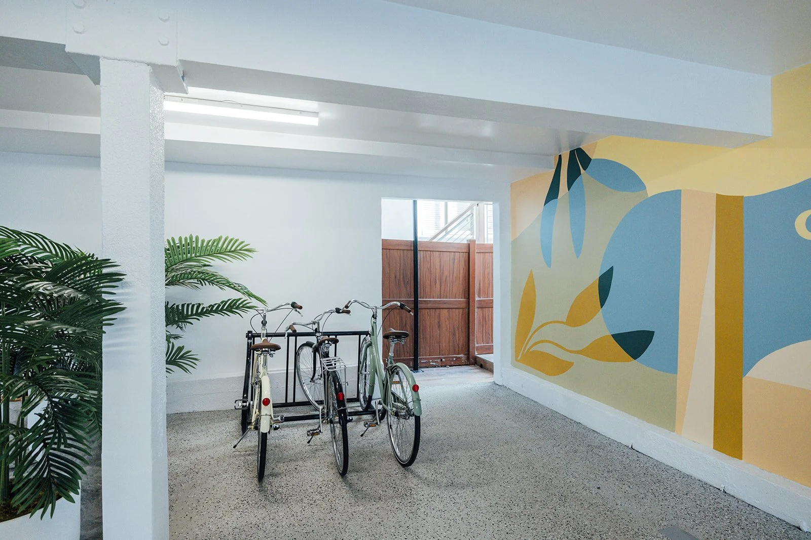 Indoor space with a decorative wall mural, bicycles parked near a wooden gate, and indoor plants.