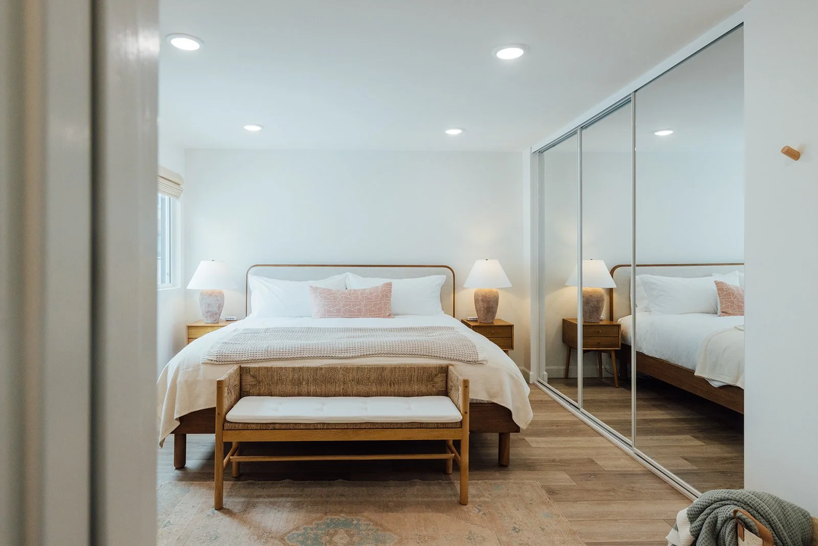 A bedroom with a bed flanked by two nightstands, each with a lamp. The bed has white linens and pink pillows. A bench is at the foot of the bed, and a large mirrored closet is on the right. The room has wooden flooring and a window on the left.