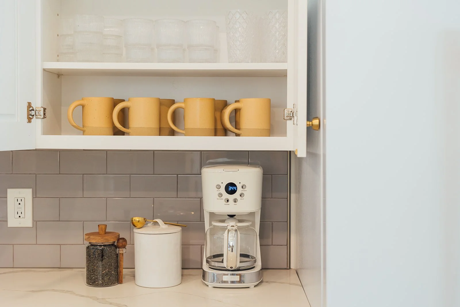Kitchen cabinet with glassware on top shelf, five yellow mugs on middle shelf, coffee maker, and containers on the counter below. Gray tiled wall with a white electrical outlet.