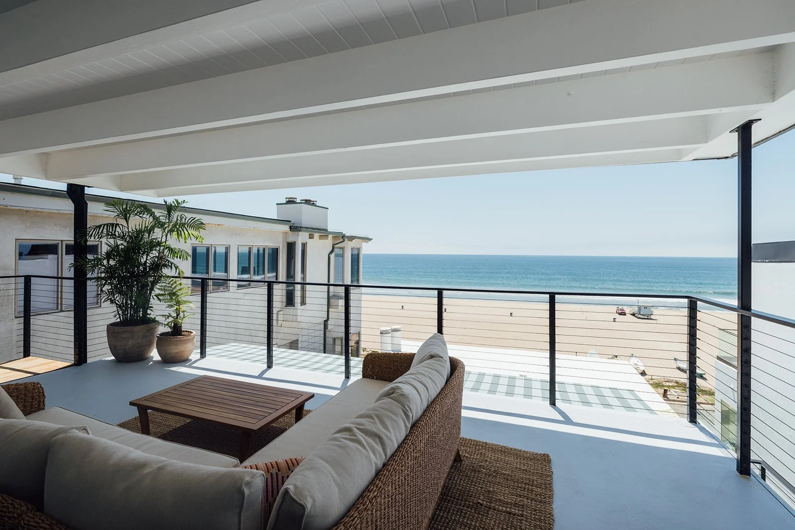 Balcony overlooking the beach and ocean, with outdoor furniture including a white sofa and a wicker chair, and potted plants under a sheltered ceiling.