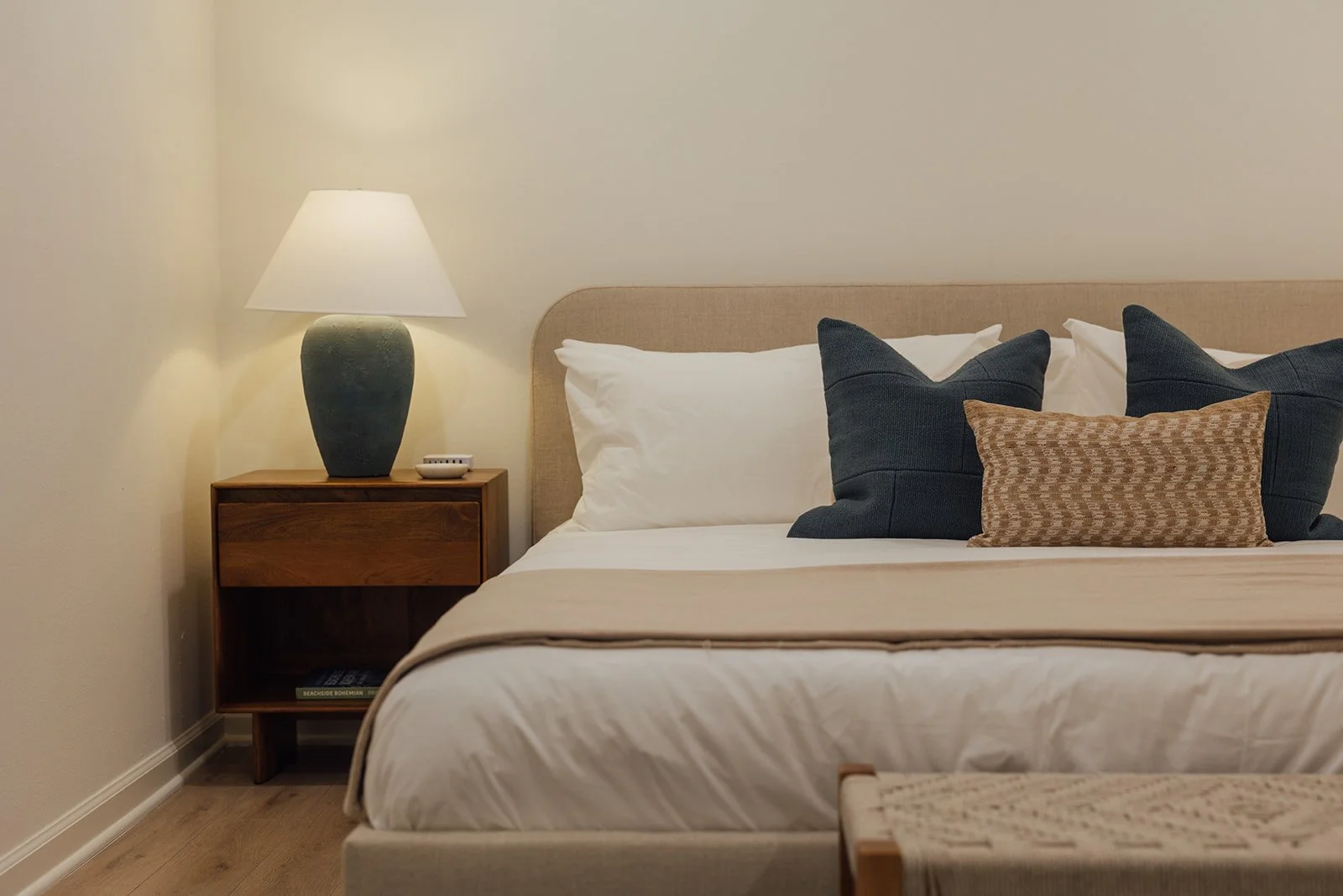 A neatly made bed with white pillows and a beige bedspread, salon-style pillows in dark gray and a small brown patterned pillow, beside a wooden nightstand with a green ceramic lamp, a small dish, and a book.