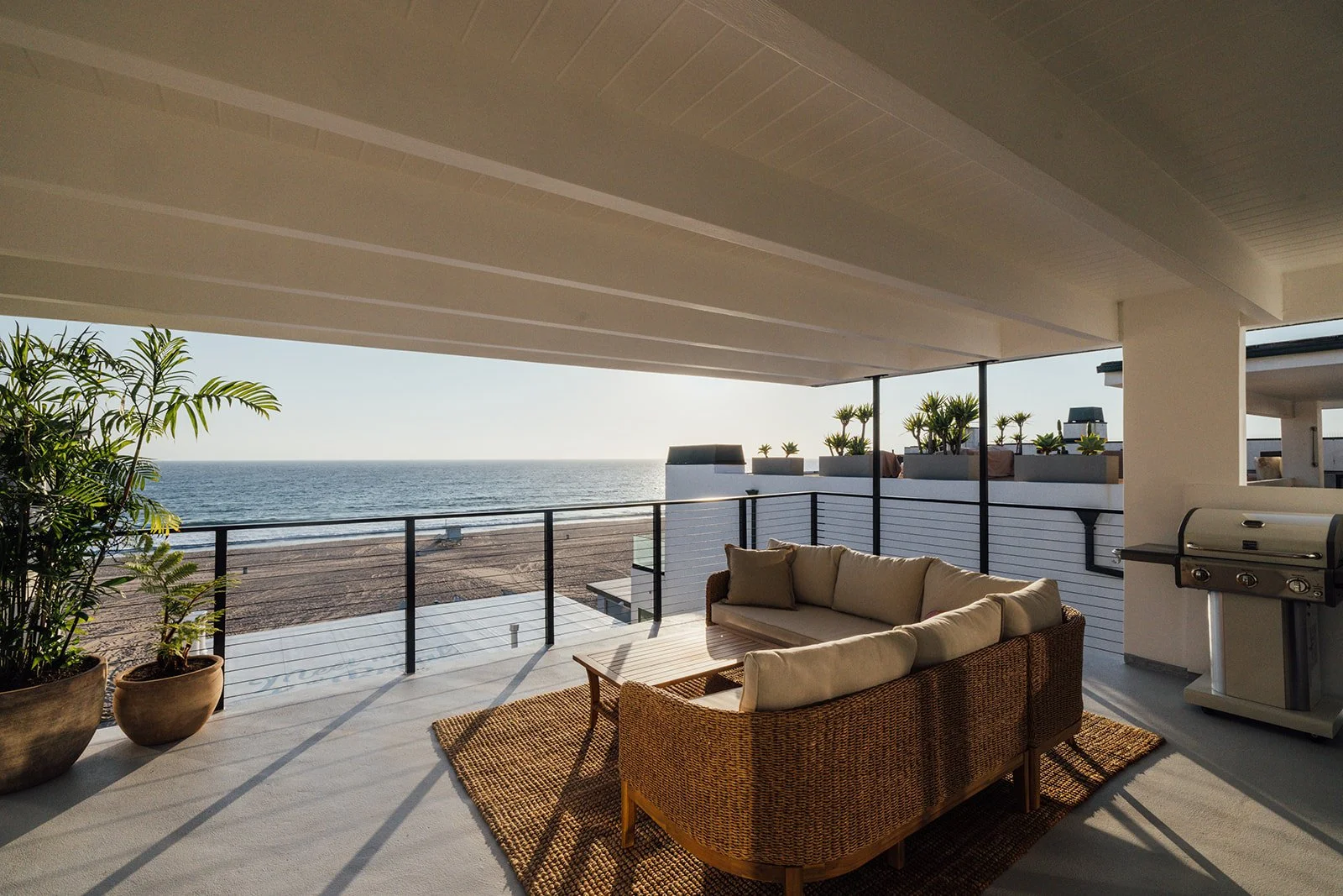 Beachside balcony with wicker sofa and coffee table overlooking the ocean, potted plants, and a BBQ grill, sunny day.