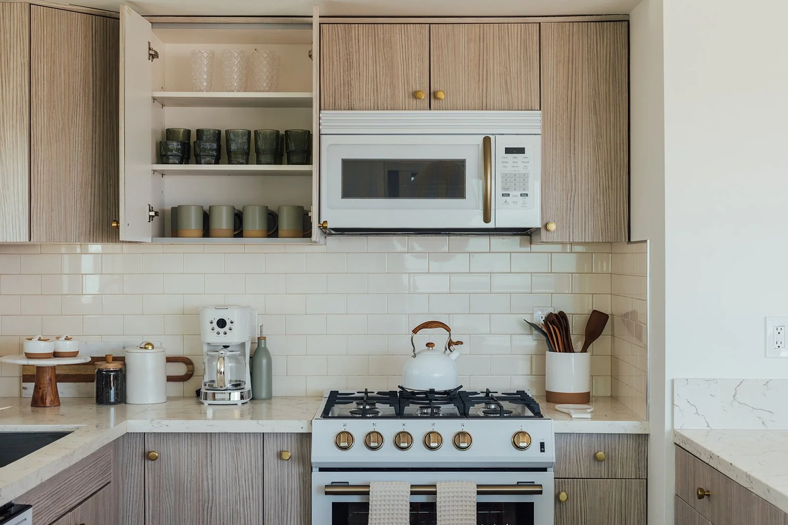 A modern kitchen with wooden cabinets, white backsplash, and white countertop. Open cabinet with glasses and coffee mugs, microwave oven above the stove, countertop appliances including a coffee maker, and various utensils.