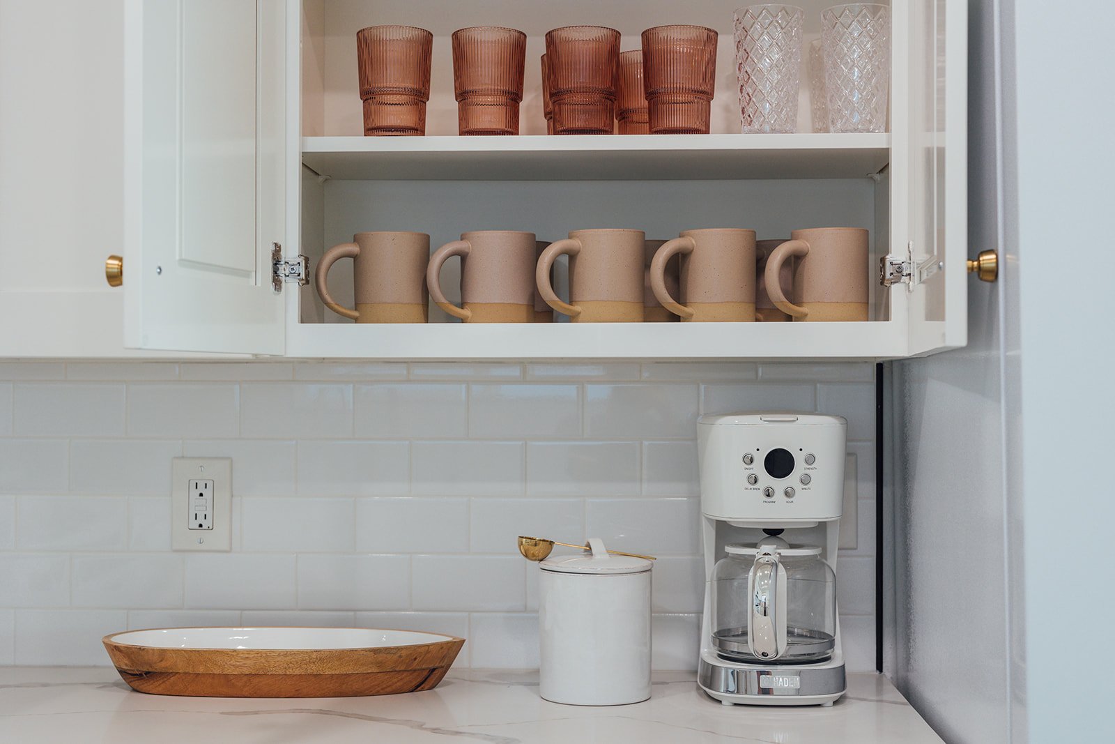 Open kitchen cabinet with four pink and beige mugs on the lower shelf and six amber and clear glasses on the upper shelf. Below the cabinet is a countertop with a wooden tray, a white container with a golden spoon, a coffee maker, and a section of white tiled wall with a power outlet.