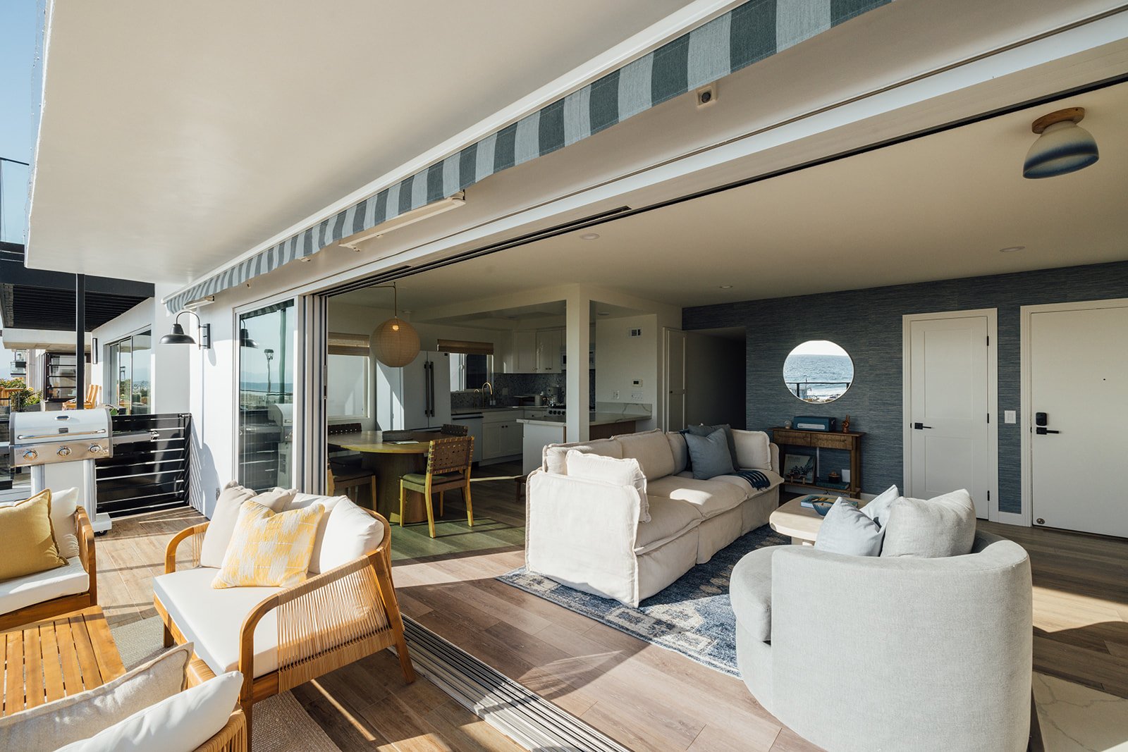 Living room with white and beige couches, a round window showing ocean view, gray textured wall, and sliding glass doors leading to a balcony with outdoor seating and a BBQ grill.