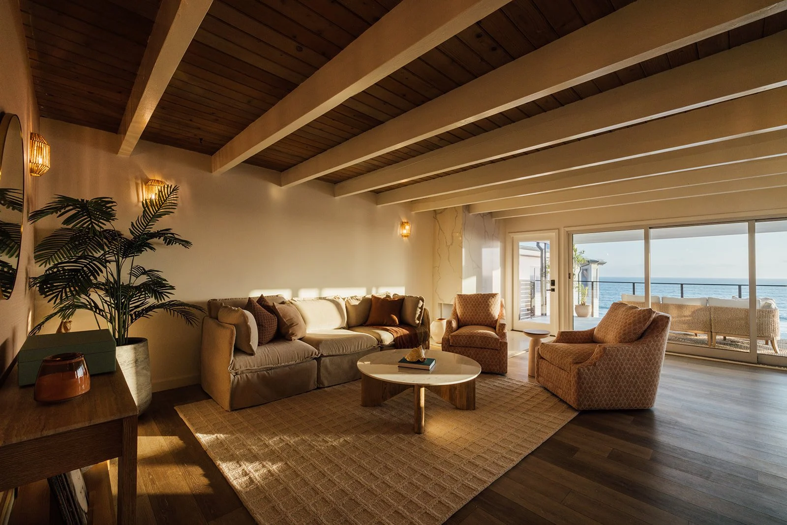 Living room with beige sofa and armchairs, wooden ceiling with exposed beams, large sliding glass door leading to balcony with ocean view, potted plant on side table, coffee table with books, and warm lighting.