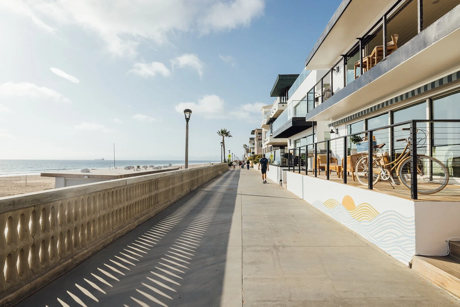 Beachside promenade with modern multi-story apartments, outdoor seating, a bicycle, and a view of the ocean with a ship in the distance.