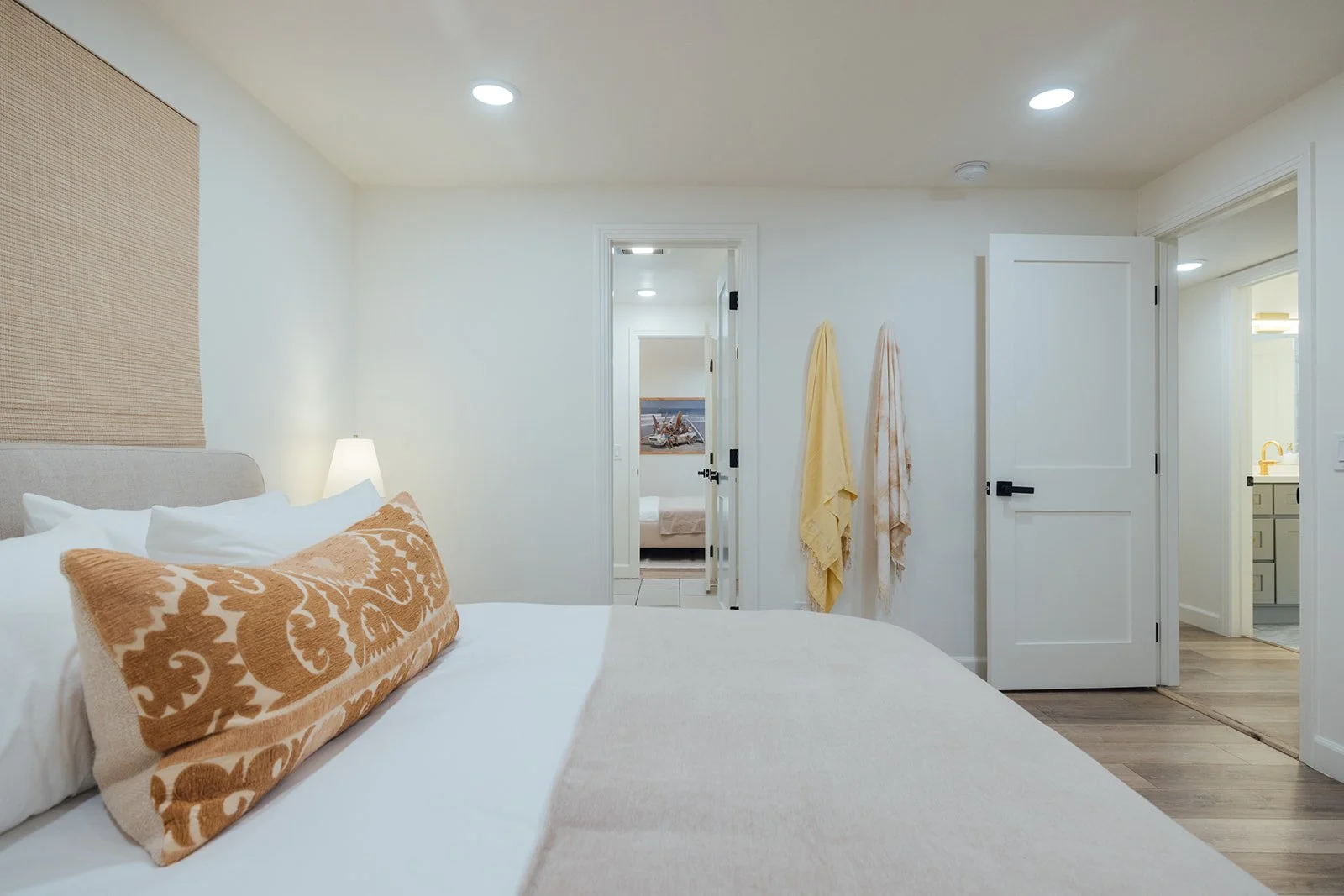 A bedroom with a bed, pillows, and a lamp, with open doors leading to a small bathroom and a room with ocean-themed artwork.