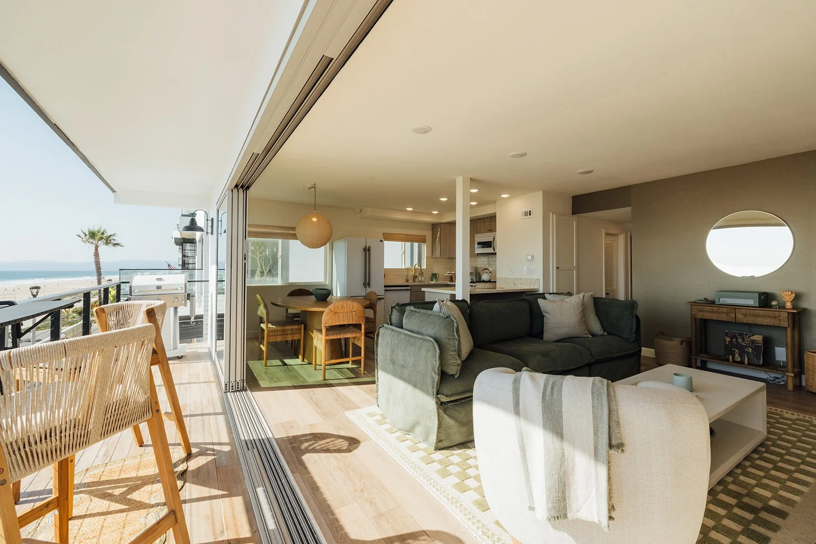 Open-concept living room and dining area with a view of the beach and ocean, featuring a balcony, a pizza oven, and modern furnishings.