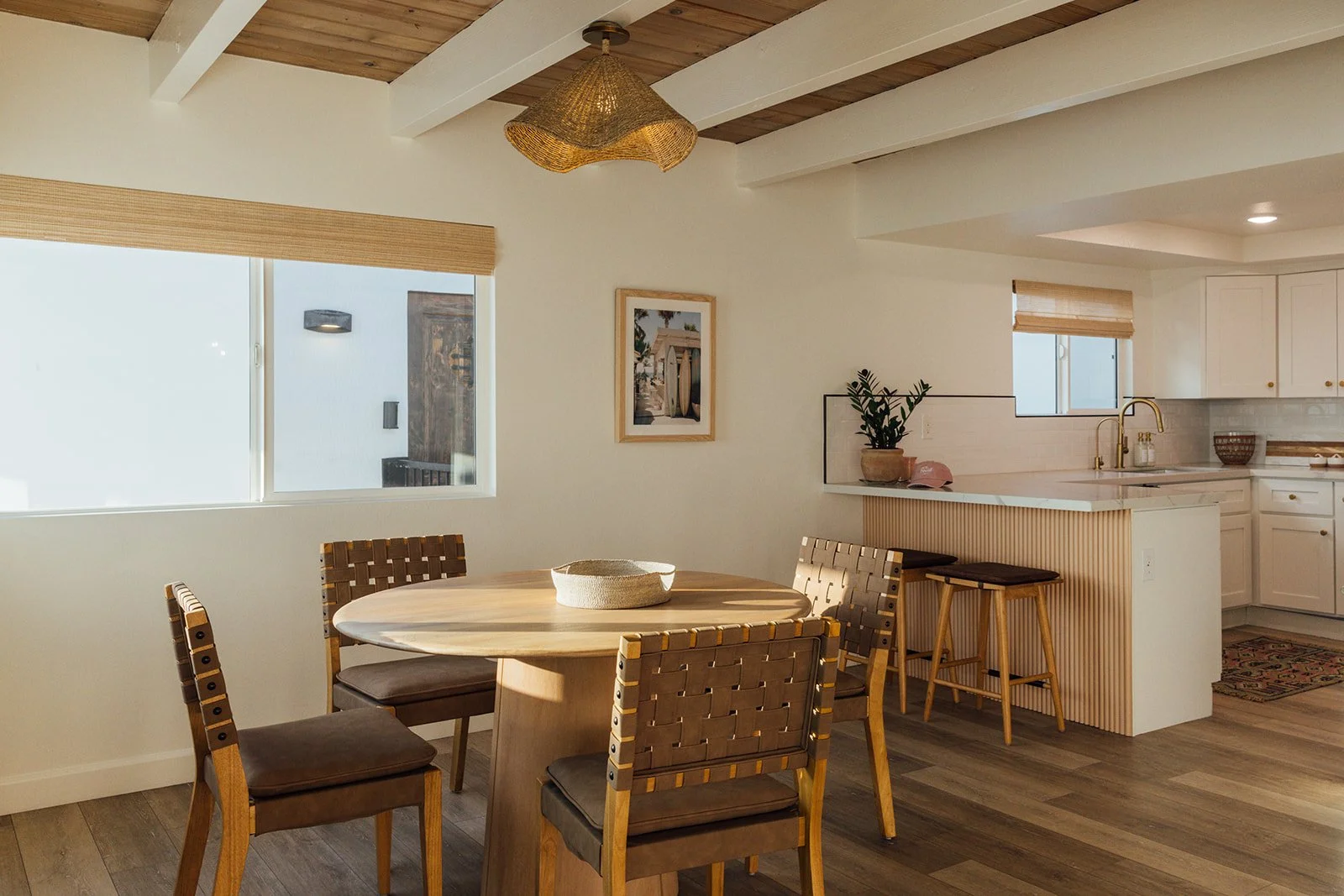 Sunlit dining area with a round wooden table surrounded by four woven chairs, located next to a kitchen with white cabinets, a marble countertop, and gold fixtures, featuring two large windows with beige blinds and a wooden ceiling with white beams.