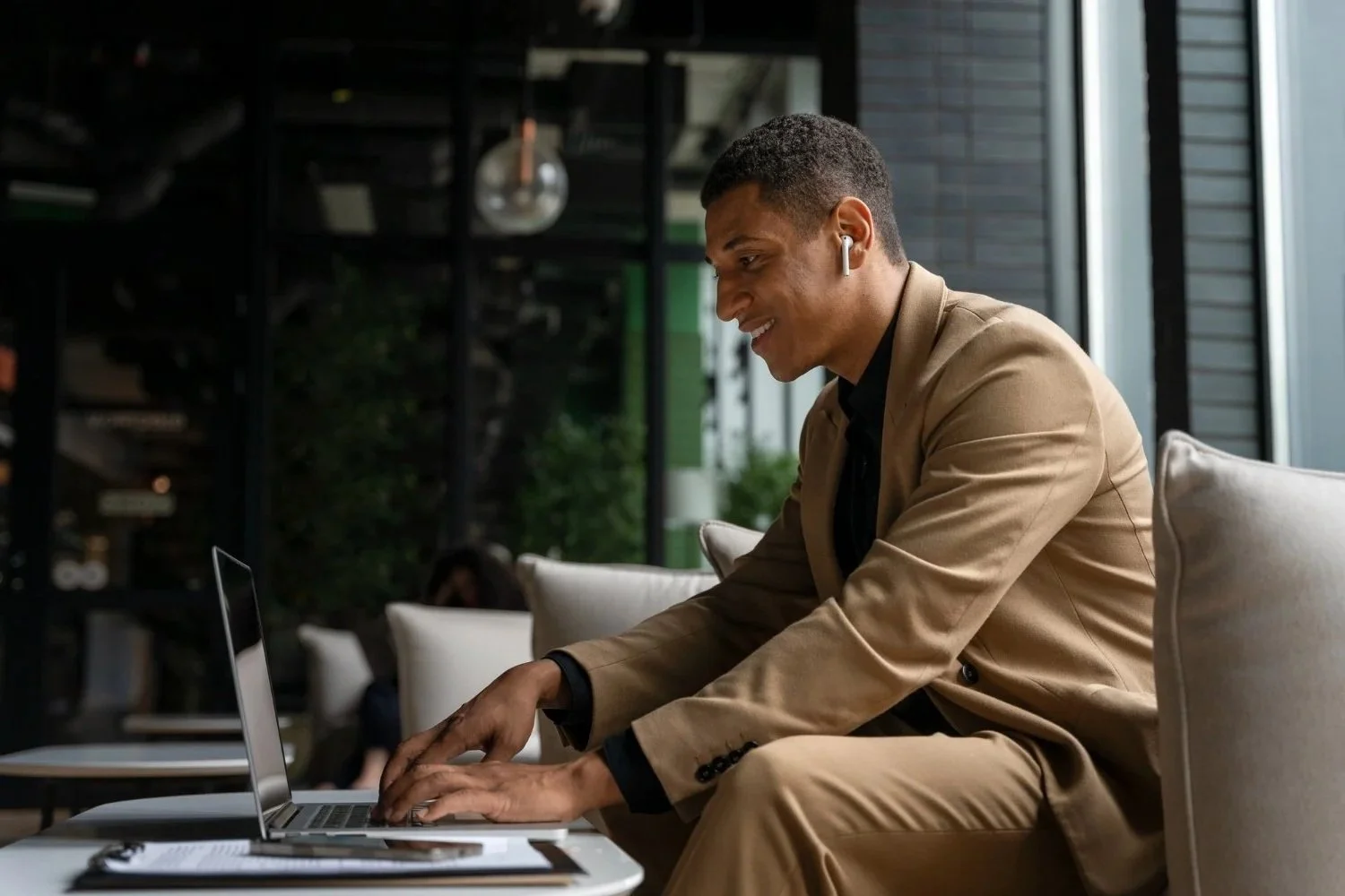 A man in a tan suit sitting on a sofa, working on a laptop with a notebook and pen nearby, wearing wireless earbuds, in a modern indoor space with large windows.