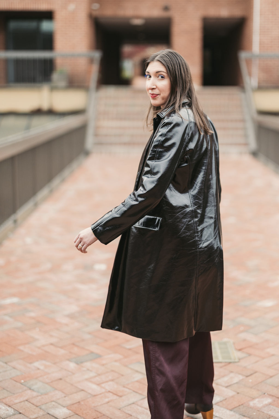 Jenn in a shiny black trench coat standing on a brick walkway, turning her head to look at the camera with a slight smile, with a brick building in the background.
