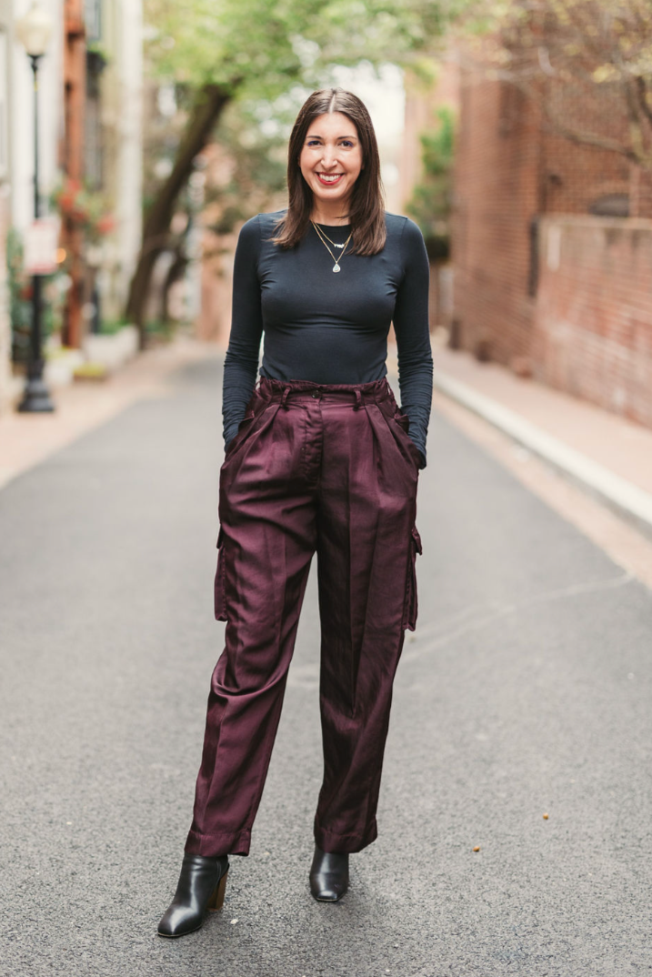 Jenn standing on a city street with trees and brick buildings in the background, wearing a dark long-sleeve top, high-waisted purple satin pants, and black ankle boots, smiling with hands in her pockets.