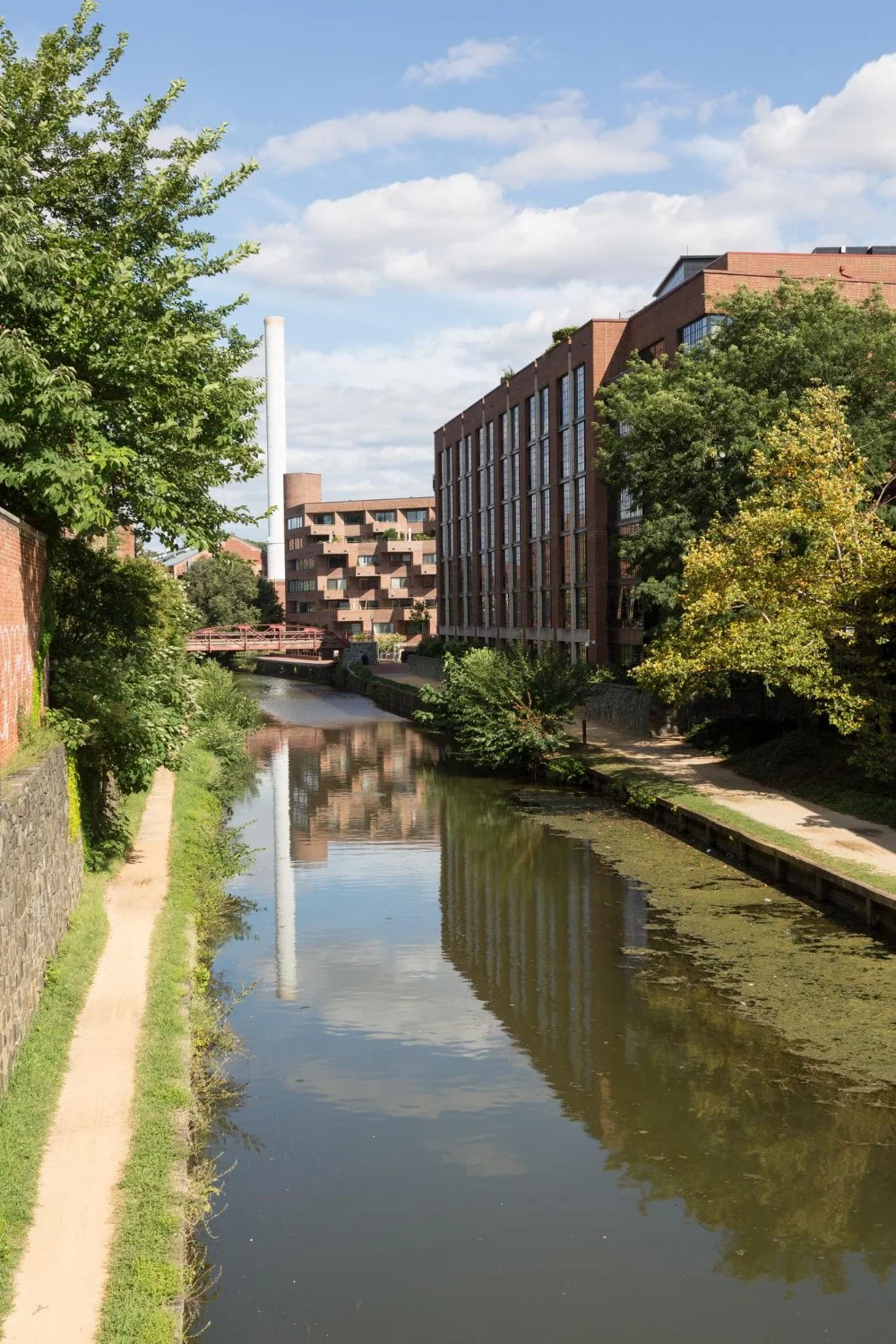 A canal in the Georgetown neighborhood of Washington, DC with trees and a walking path on one side, modern brick buildings on the other, under a partly cloudy sky.