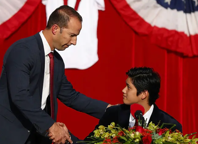 A man in a dark suit and red tie shakes hands with a young man seated at a table with a bouquet of flowers and a microphone, in front of red curtains and patriotic bunting.