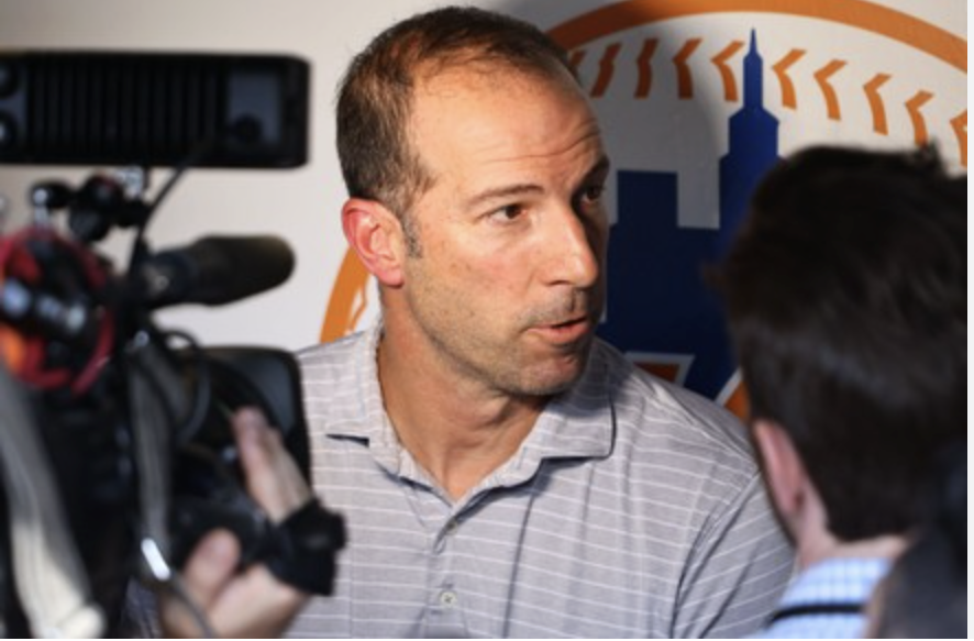 A man being interviewed by a reporter with a camera recording the interaction, in front of a New York Mets logo.