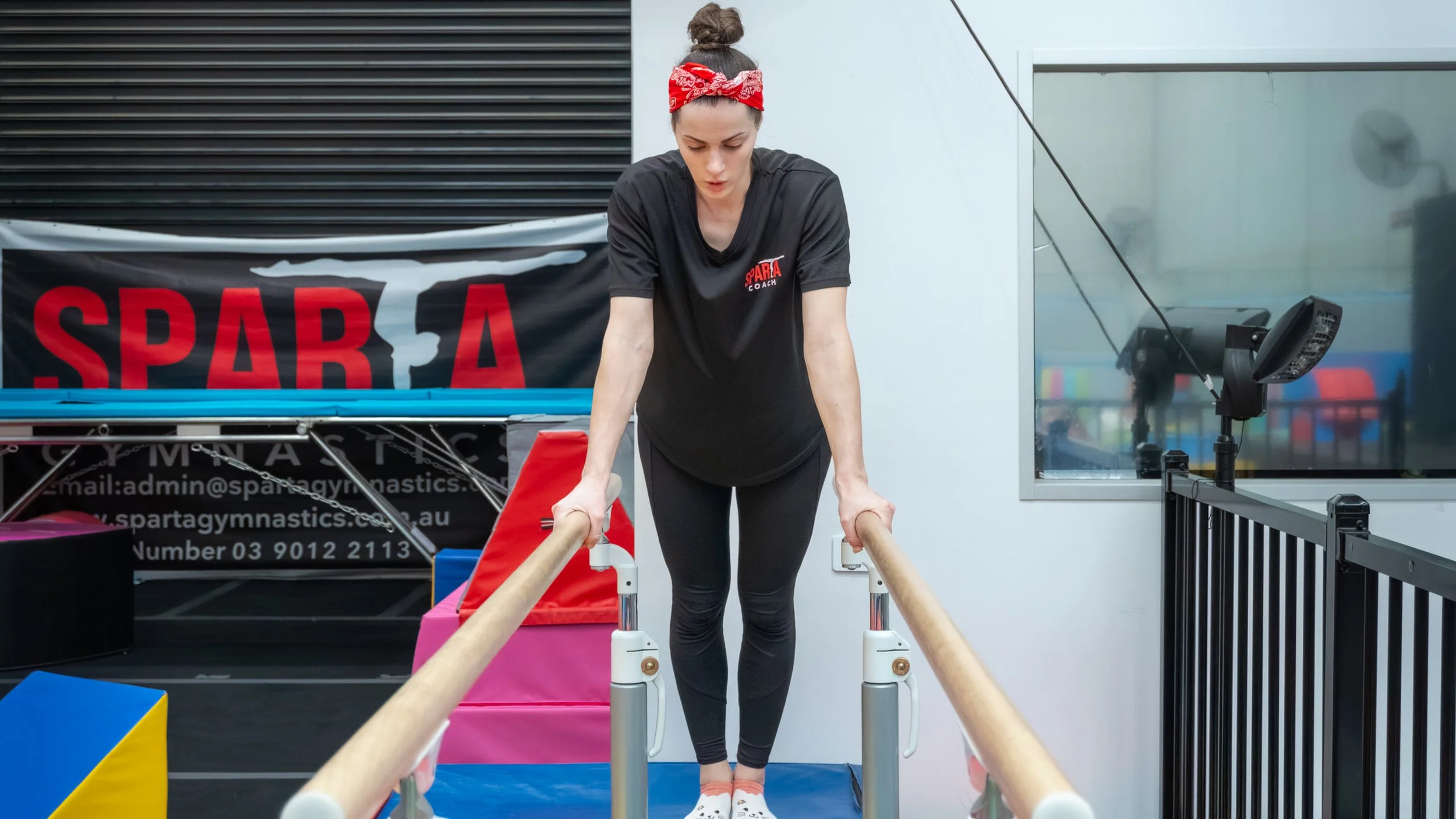 A woman doing a gymnastics balance exercise on parallel bars in a gym, wearing a black t-shirt with red and white logo, black leggings, and a red bandana in her hair.