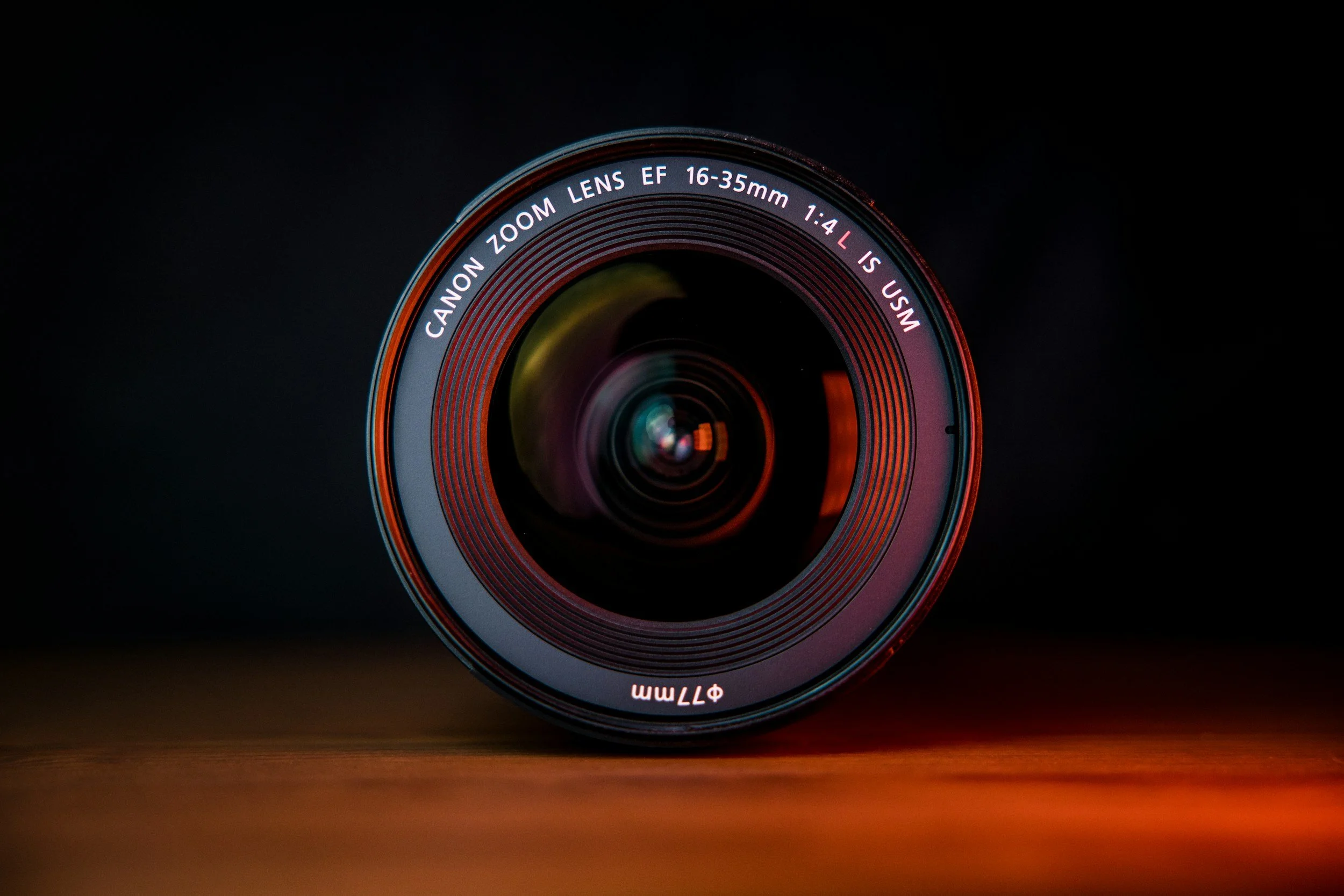 A Canon zoom lens with a focus range 16-35mm, lying on a wooden surface with a dark background.
