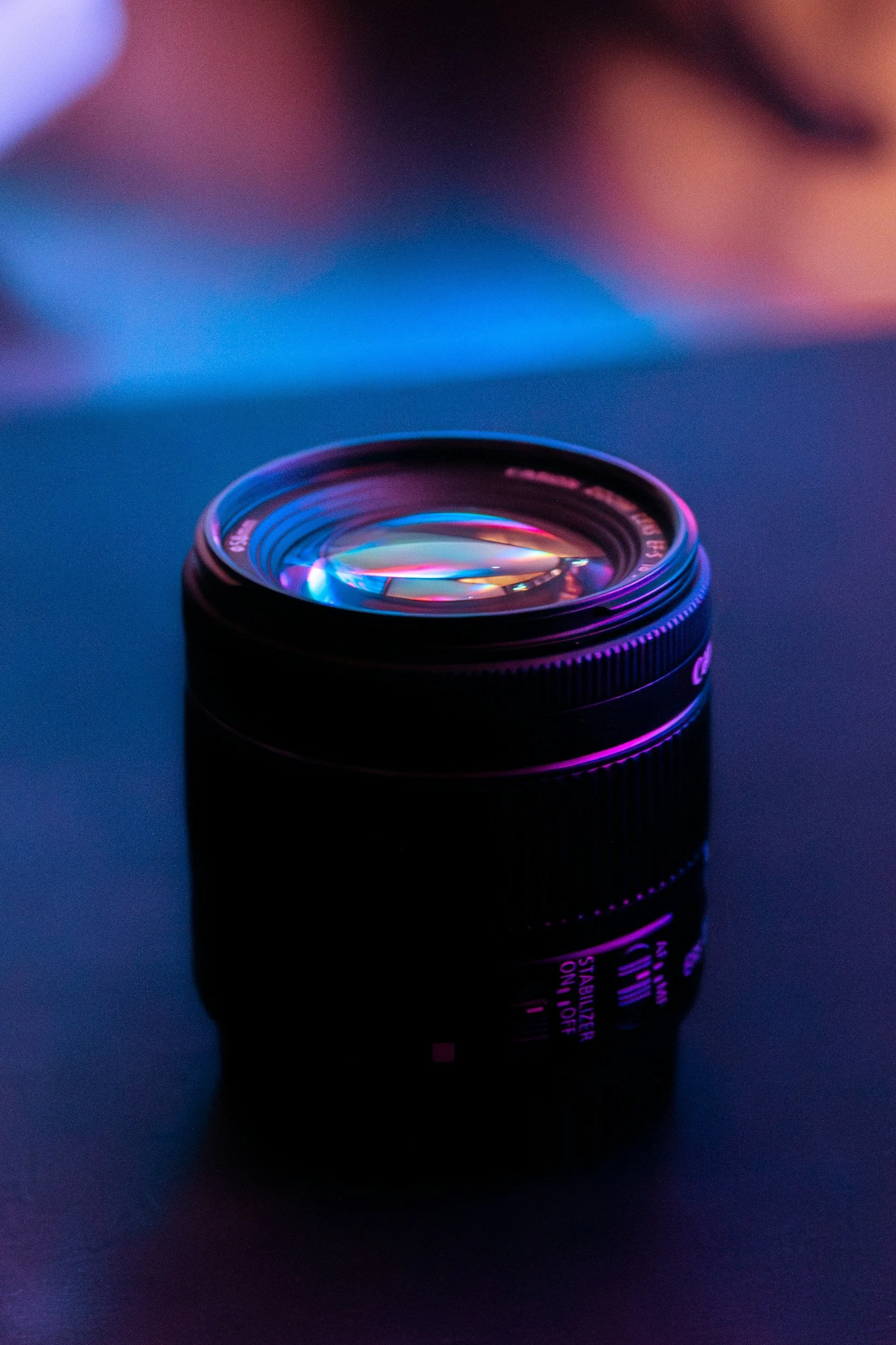 A camera lens with a colorful lens filter reflects light under purple and blue lighting.
