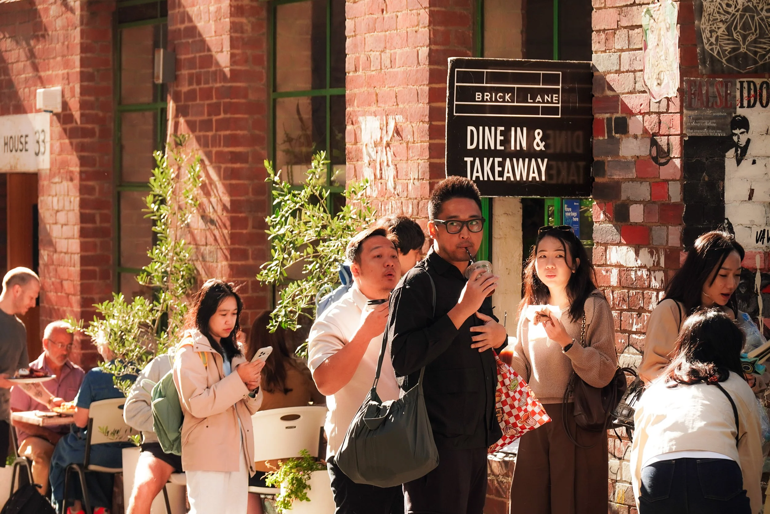 People standing outside a brick cafe sign reading 'BRICK LANE DINE IN & TAKEAWAY' on a sunny day.