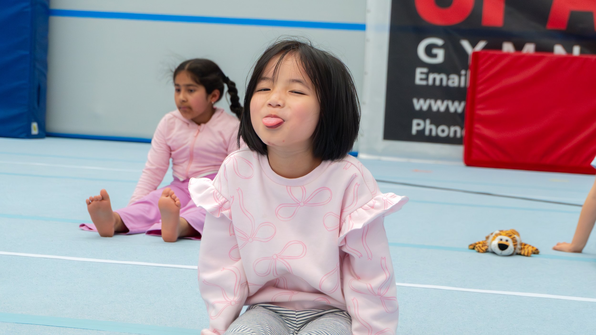 A young girl with short black hair sticking out her tongue, sitting on the blue floor of a gym, with a girl in pink outfit sitting behind her and a small plush tiger toy nearby.