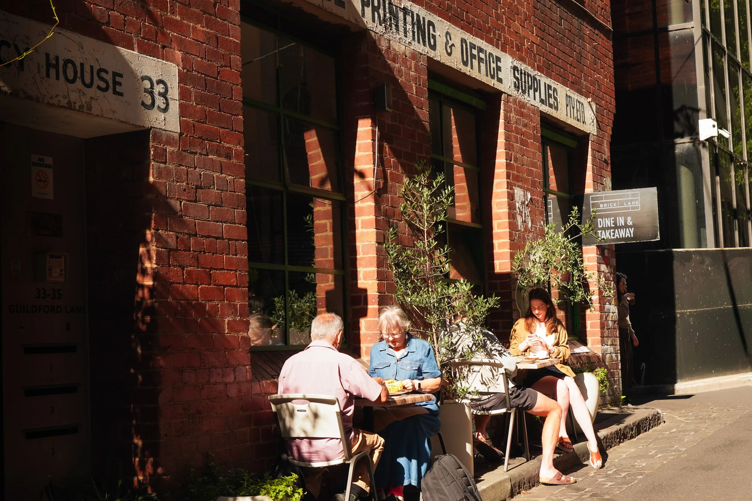 People sitting outside a brick building with windows, a sign that says dine in and takeaway, and street signs indicating the location as Guildford Lane.