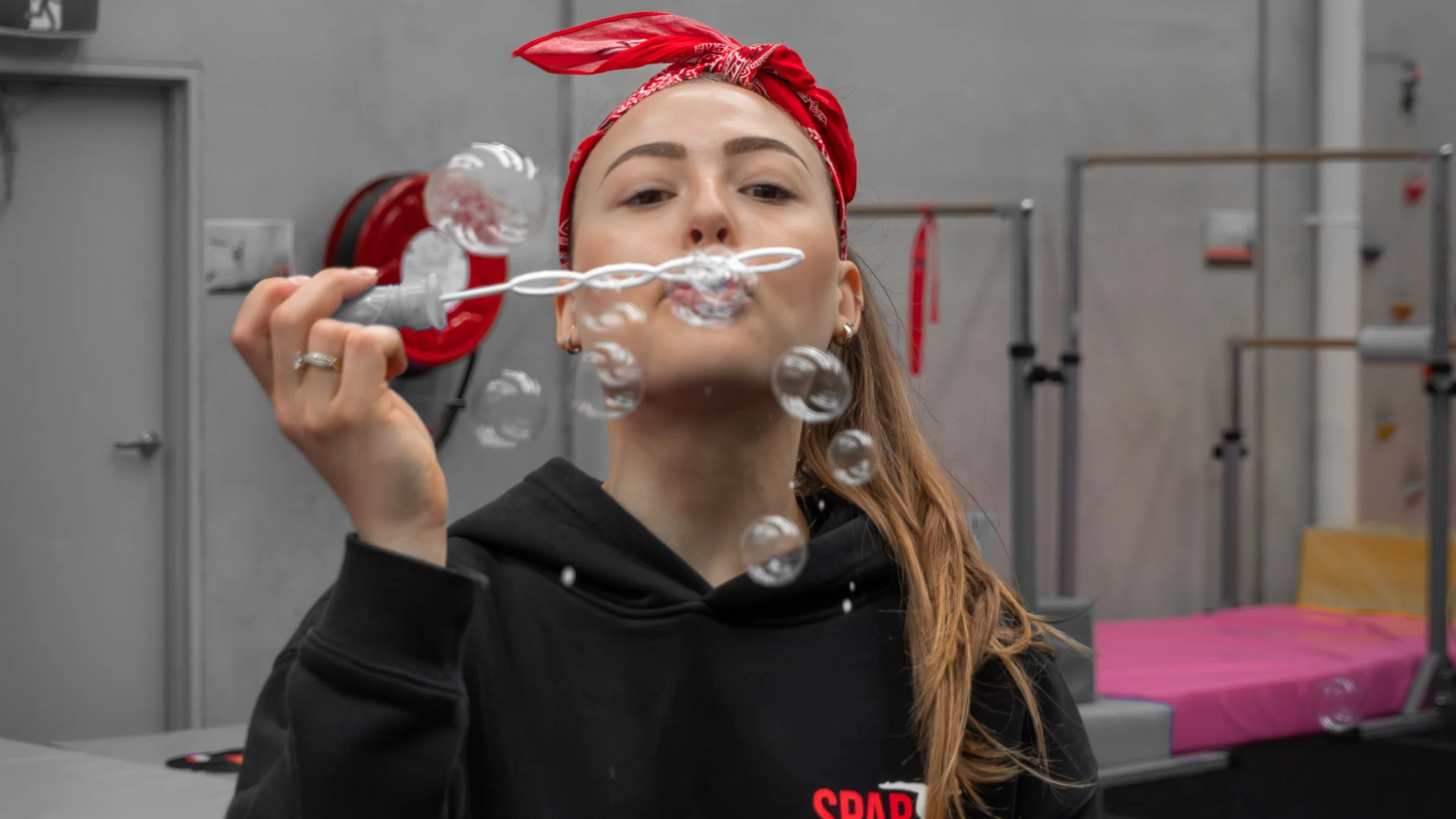 A woman with a red bandana blowing bubbles in a gym or training facility.