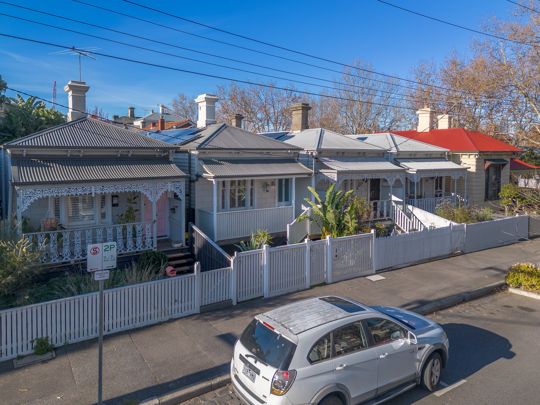 A row of Victorian-style houses with colorful roofs, white fences, and small front gardens, parked silver car in foreground, street parking sign, clear blue sky, leafless trees in background.