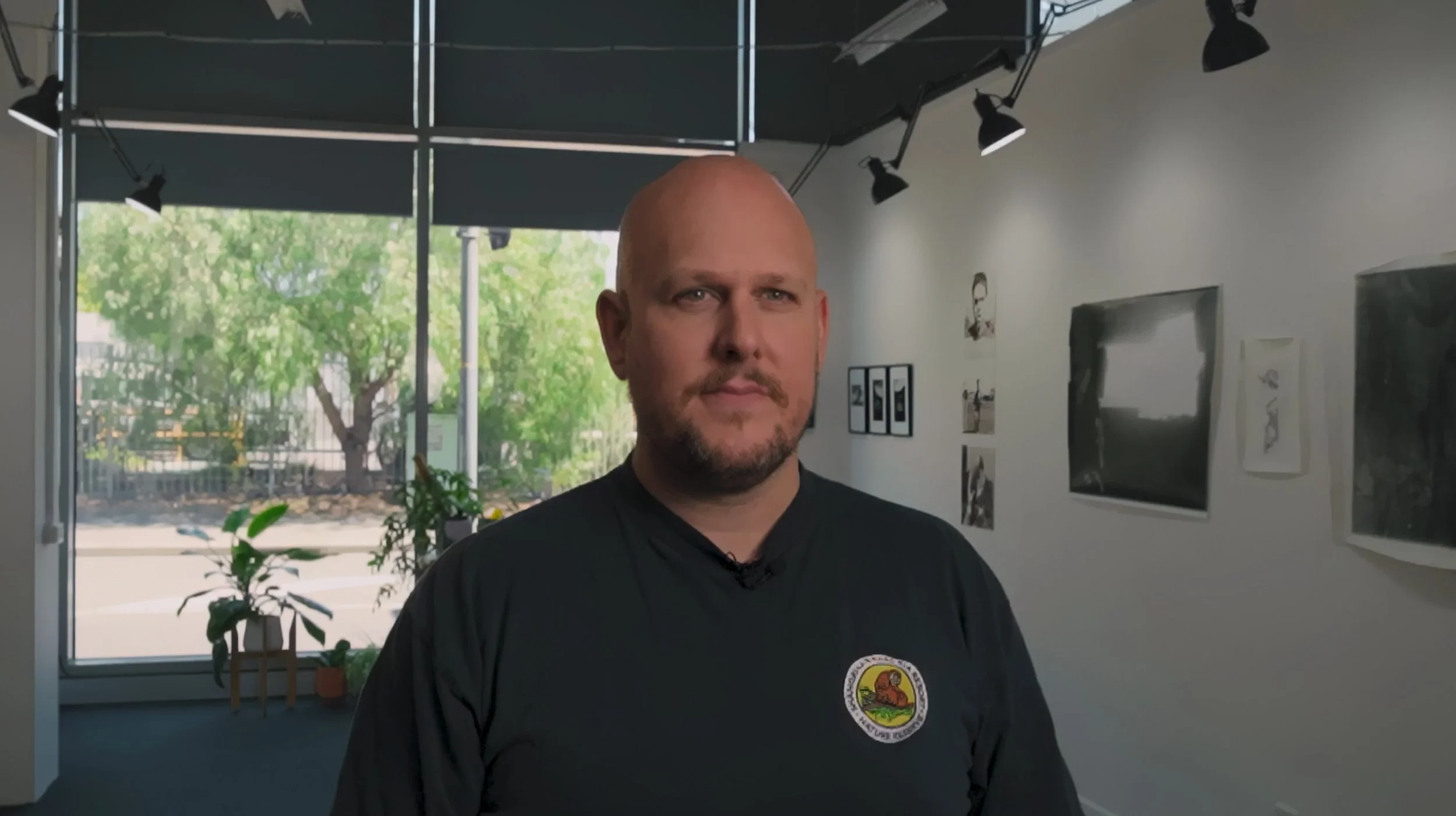 Bald man with a beard standing inside a room with artwork on the walls and a large window with trees outside.