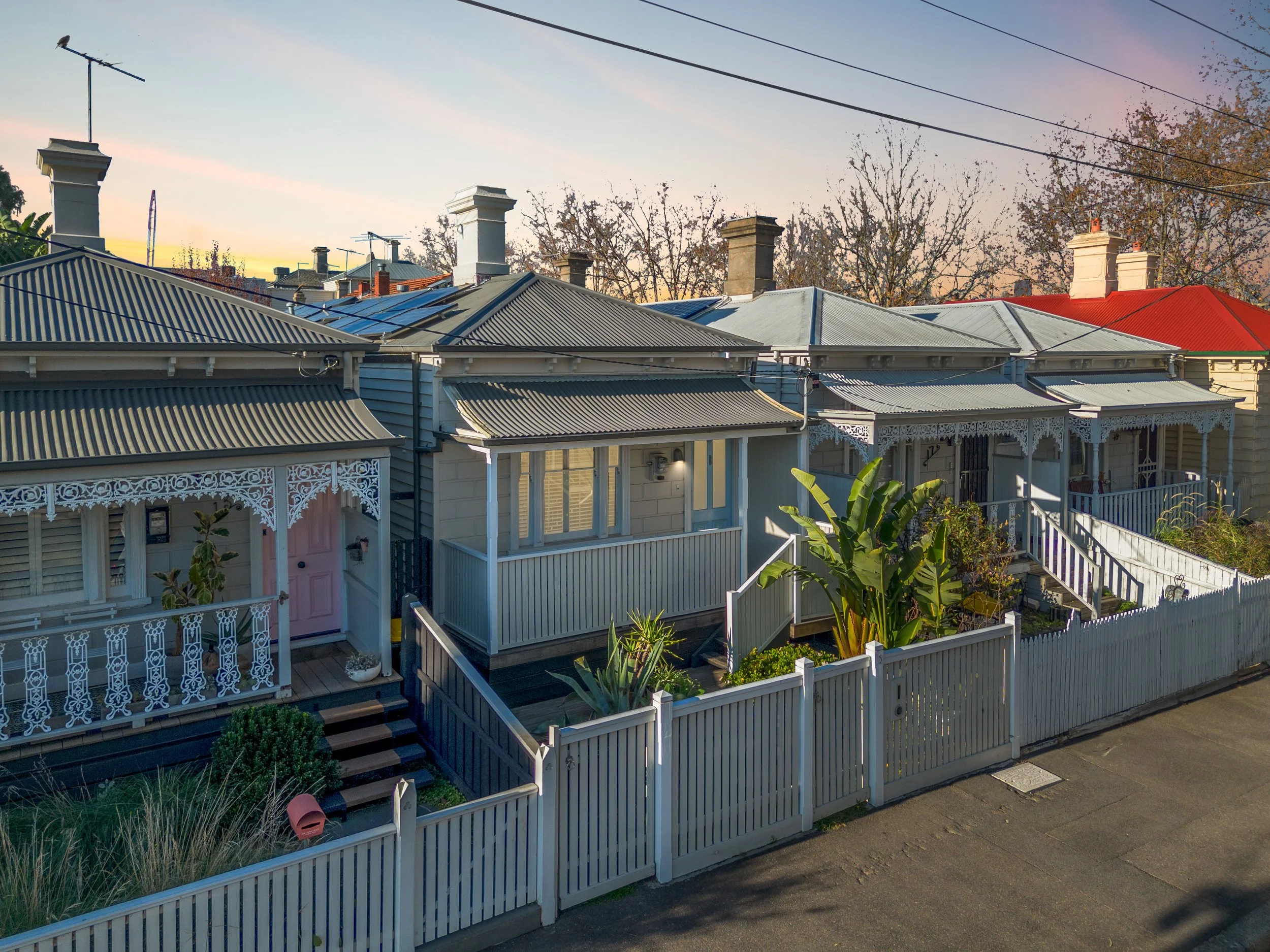 A row of Victorian-style houses with ornate porch trims, metal roofs, and small front gardens enclosed by white picket fences, under a sunset sky.