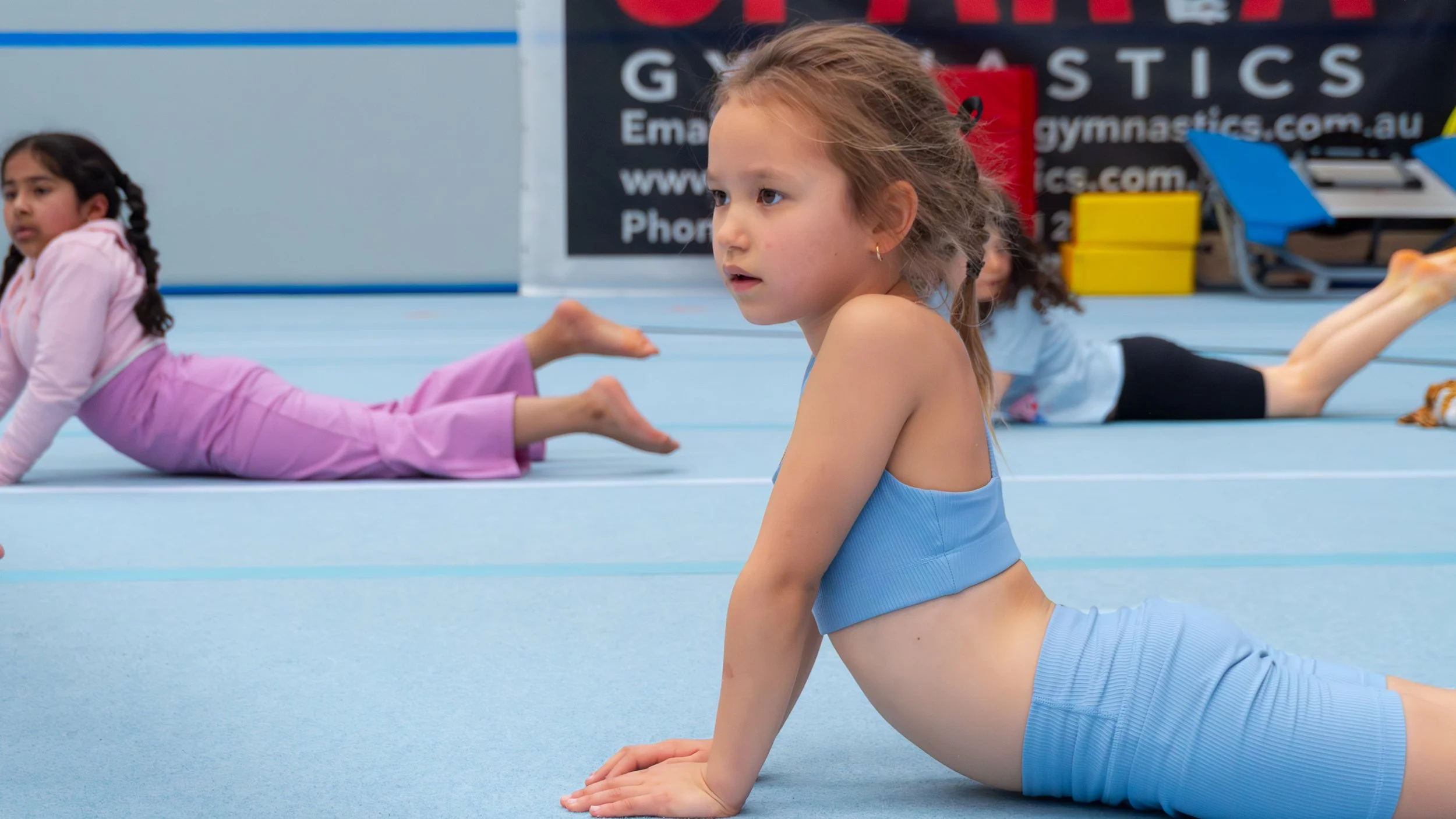 Young girls practicing gymnastics, with one girl in the foreground doing the cobra pose on the floor and two others in the background stretching on a gymnasium mat.