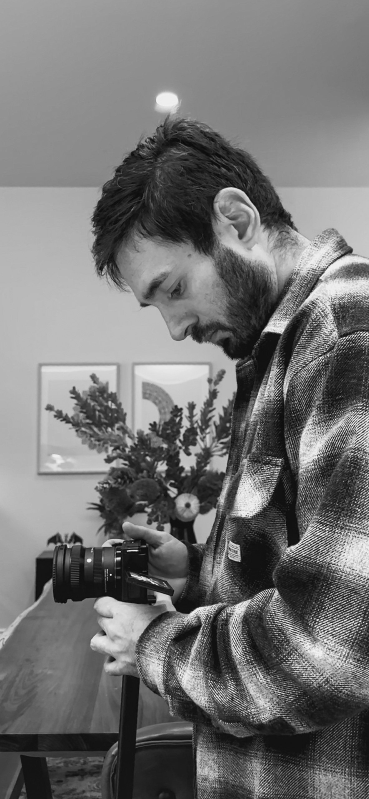 A young man with dark hair and a beard, wearing a plaid shirt, is looking down at a camera he is holding in his hands in a room with framed pictures on the wall and a large floral arrangement in the background.