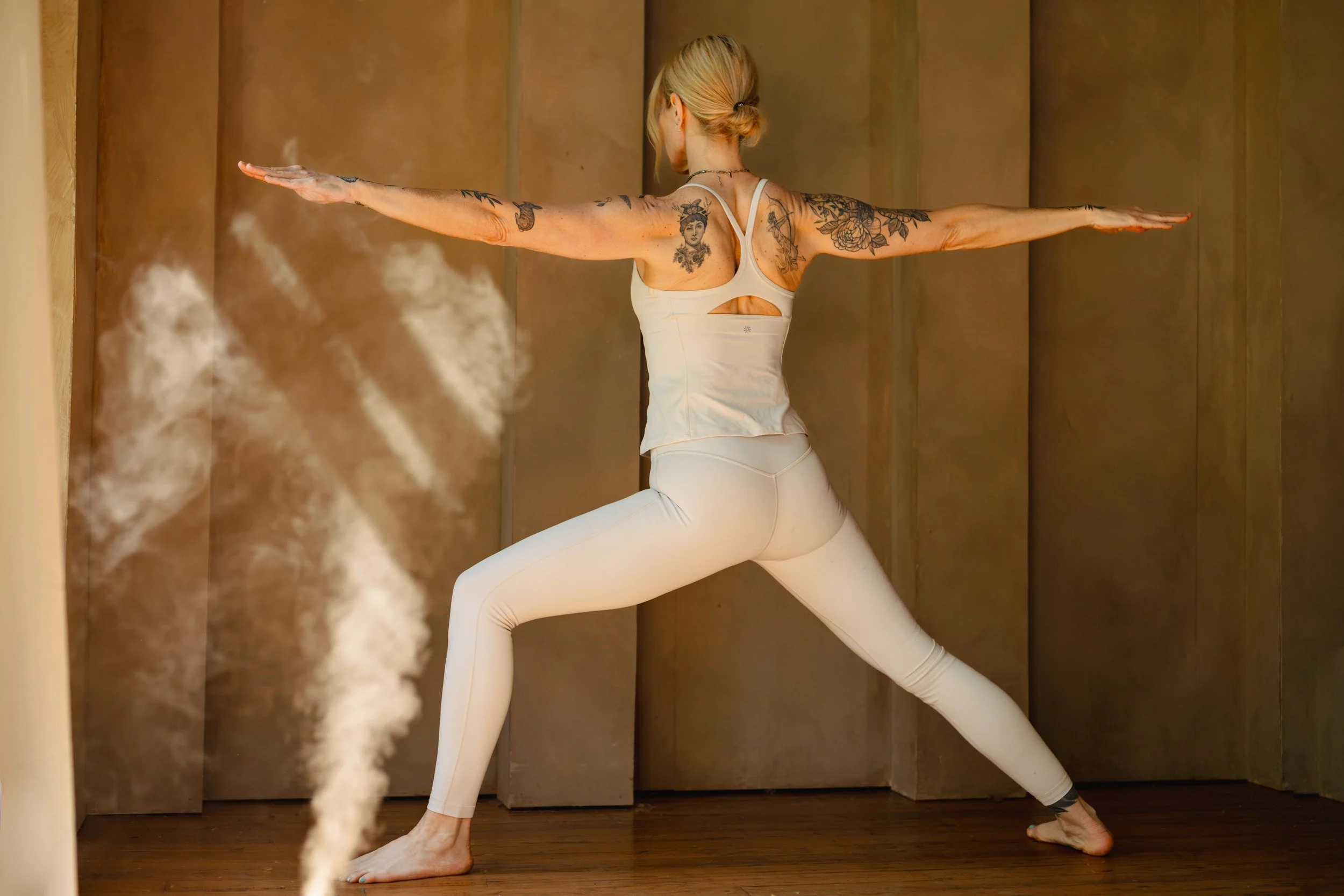 A woman practicing yoga in a side lunge position, with her arms extended outward, indoors with wooden walls and floor, and steam or vapor around her.