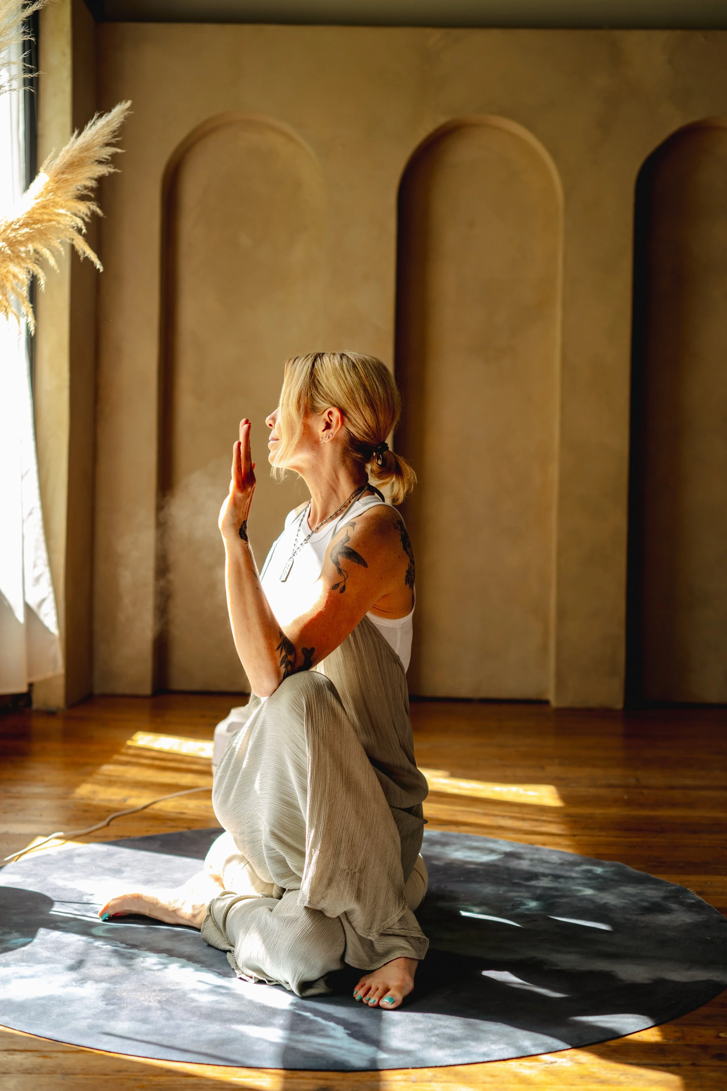 A woman with blonde hair, tattoos, and a white top is practicing yoga in a sunlit room, kneeling on a black mat with her eyes closed and her hand near her face, seated in a half-lotus pose.