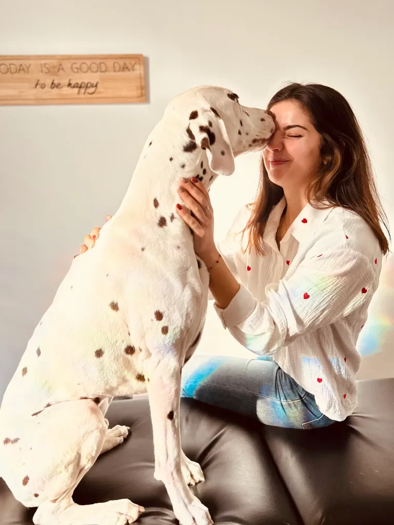 Une femme souriante en chemise blanche avec des petits cœurs rouges, assise sur un canapé, reçoit un bisou affectueux d'un grand chien Dalmatien blanc avec des taches noires.