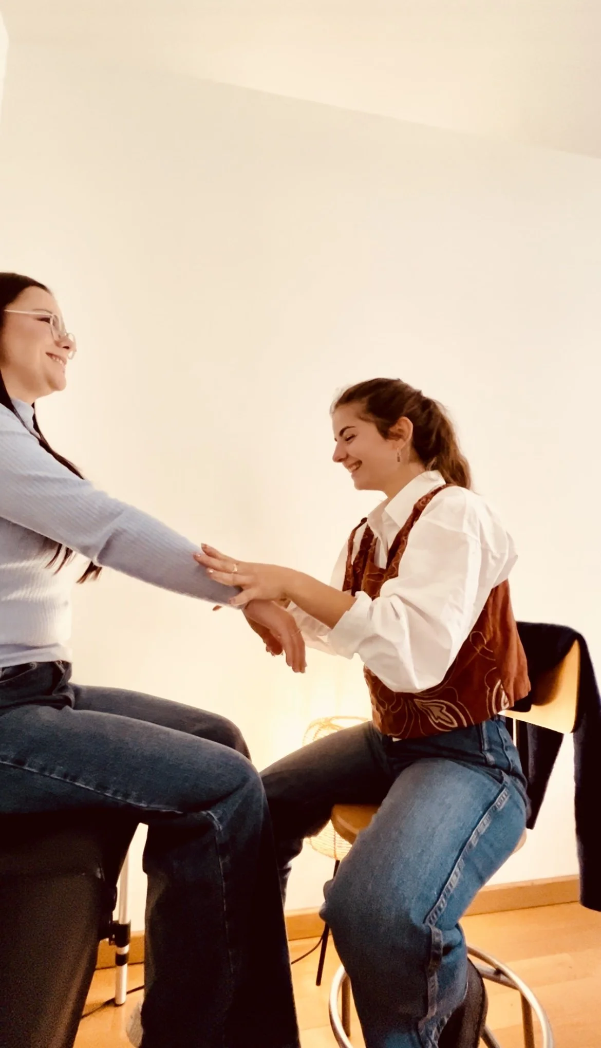 Deux femmes souriantes, une assise et l'autre à genoux, se tenant par les mains dans une pièce avec un fond blanc et un éclairage chaud.