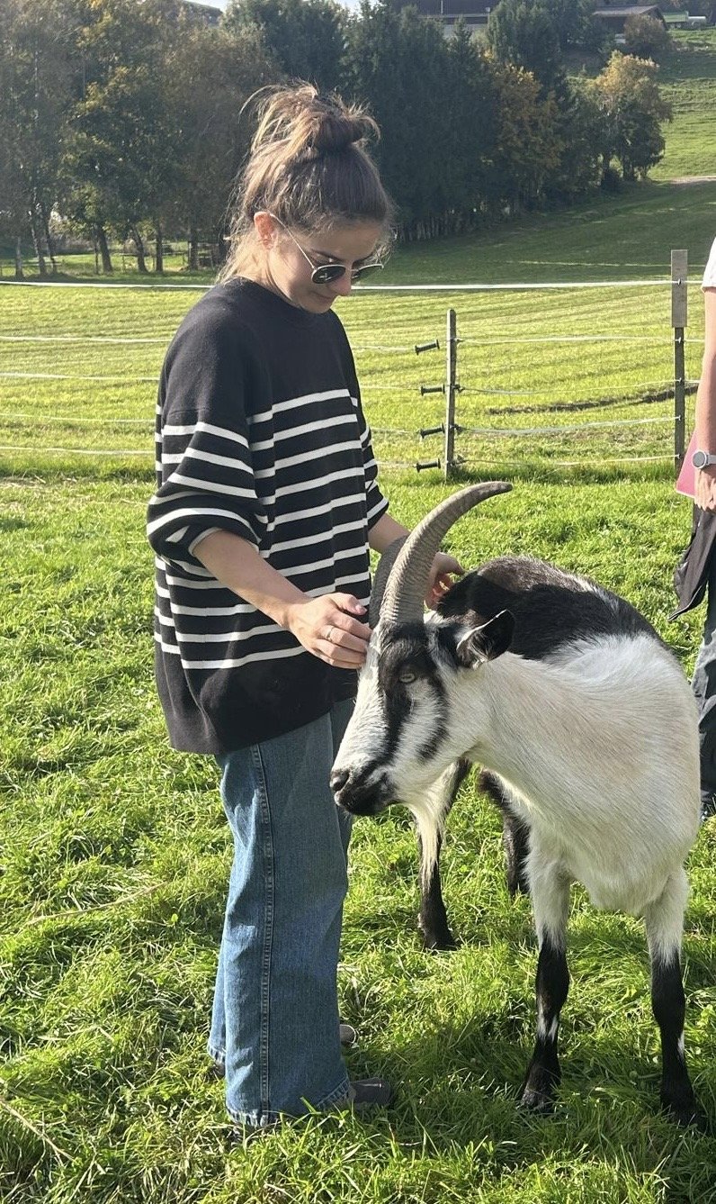 Une femme portant un sweat à rayures noir et blanc et des lunettes de soleil caresse une cabra dans un champ vert en plein air, avec un paysage de collines et d'arbres en arrière-plan.