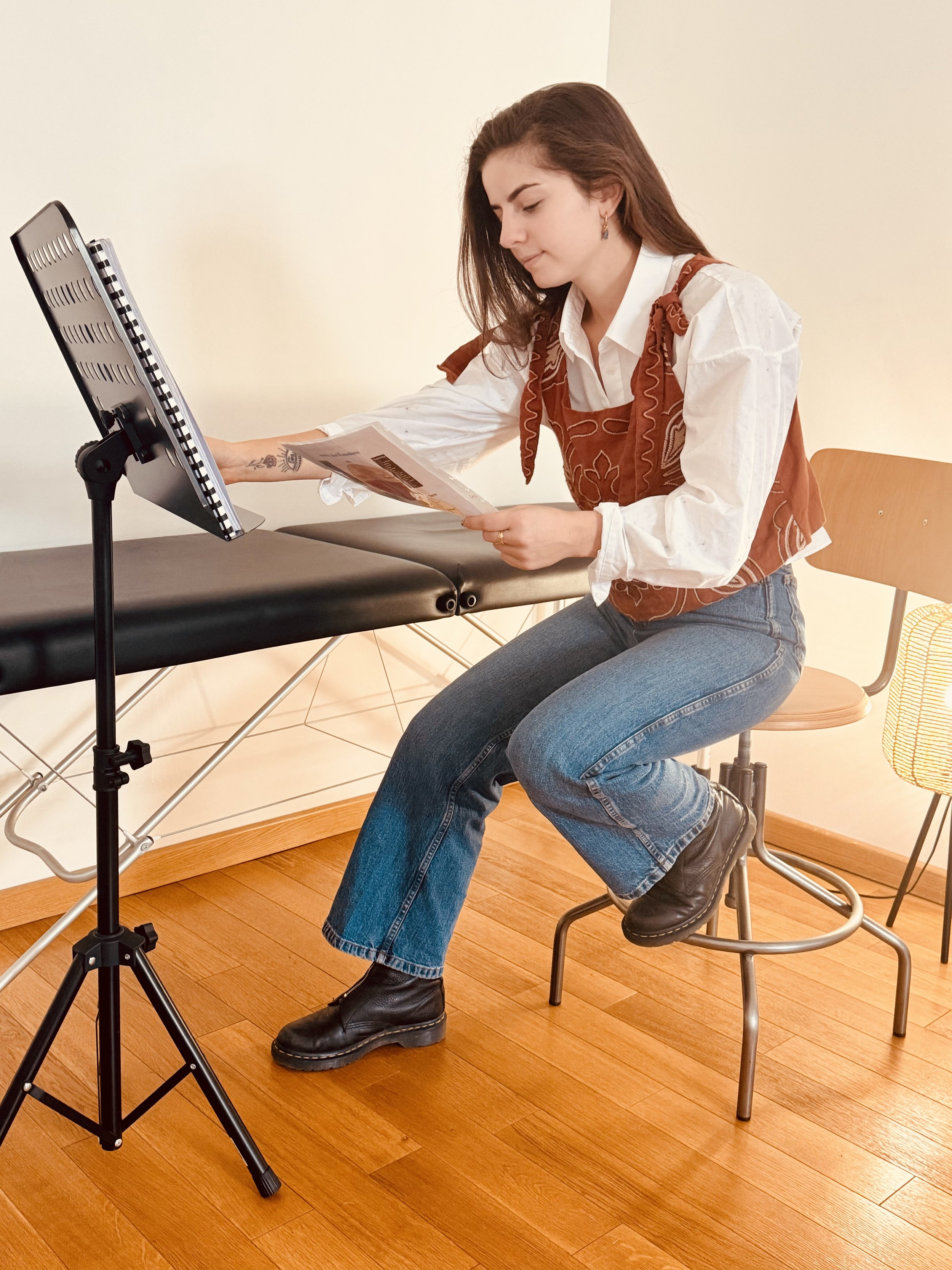 Jeune femme assise sur une chaise en studio, regardant un script ou une brochure, avec une table de massage en arrière-plan et une lampe à côté.