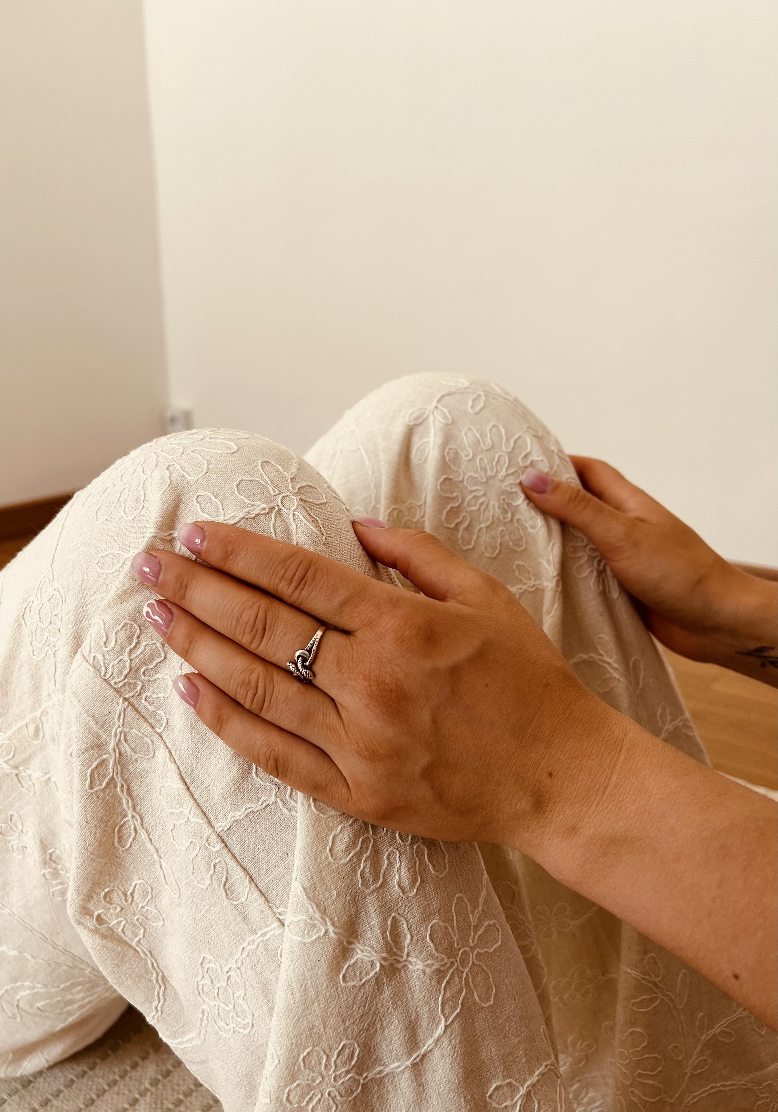 Main d'une femme posée sur son genou, portant une alliance en forme de tige avec un nœud, vêtue d'une jupe blanche avec broderies florales. Fond neutre et sol en bois.