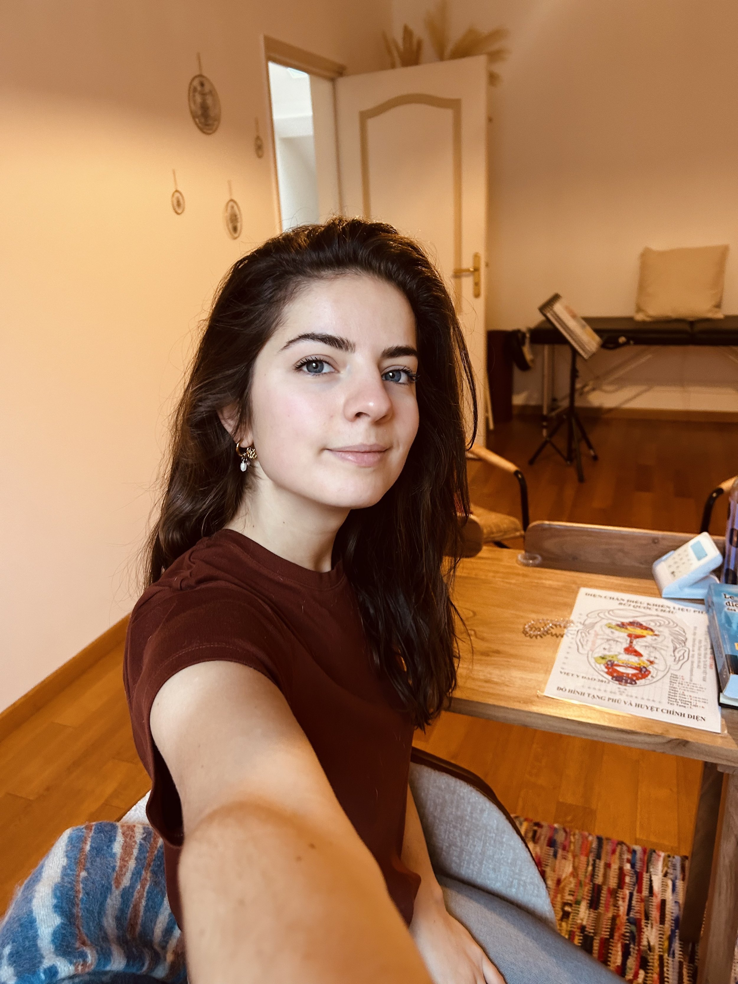 Jeune femme aux cheveux bruns, portant un t-shirt marron, prenant un selfie dans une pièce avec une chaise, une table avec des livres et un téléphone fixe.