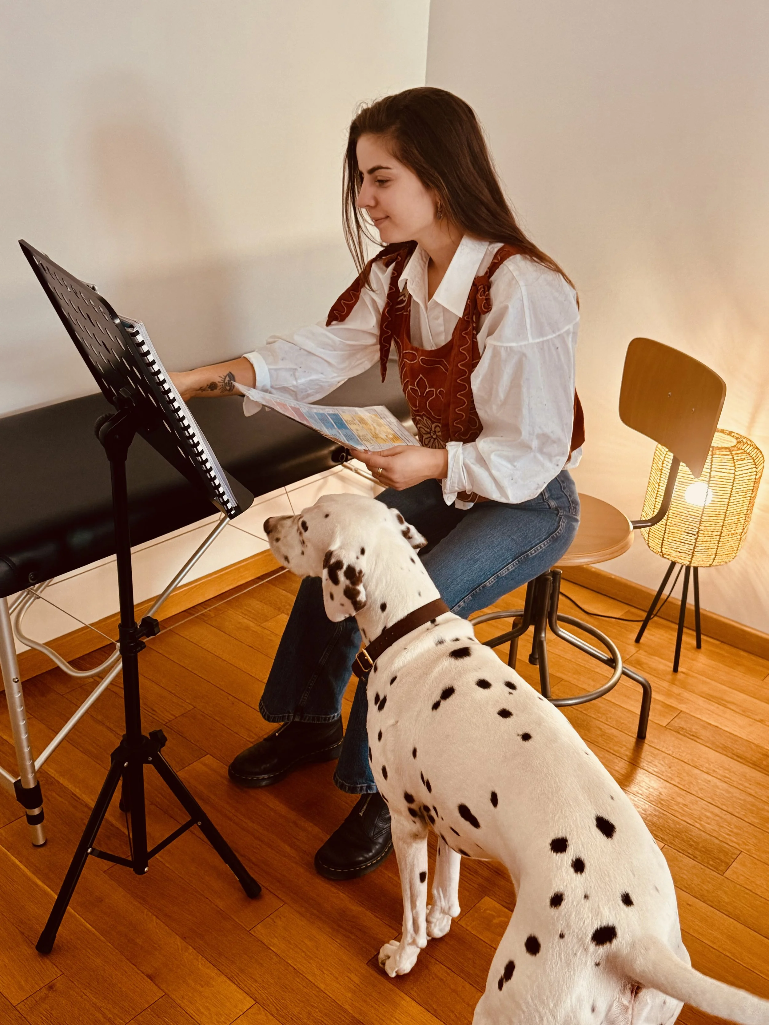 Une femme assise sur une chaise, regardant une feuille ou un menu, avec un Dalmatien assis à ses côtés. La pièce est chaleureuse, avec un lampadaire allumé, un sol en bois et un mur blanc.