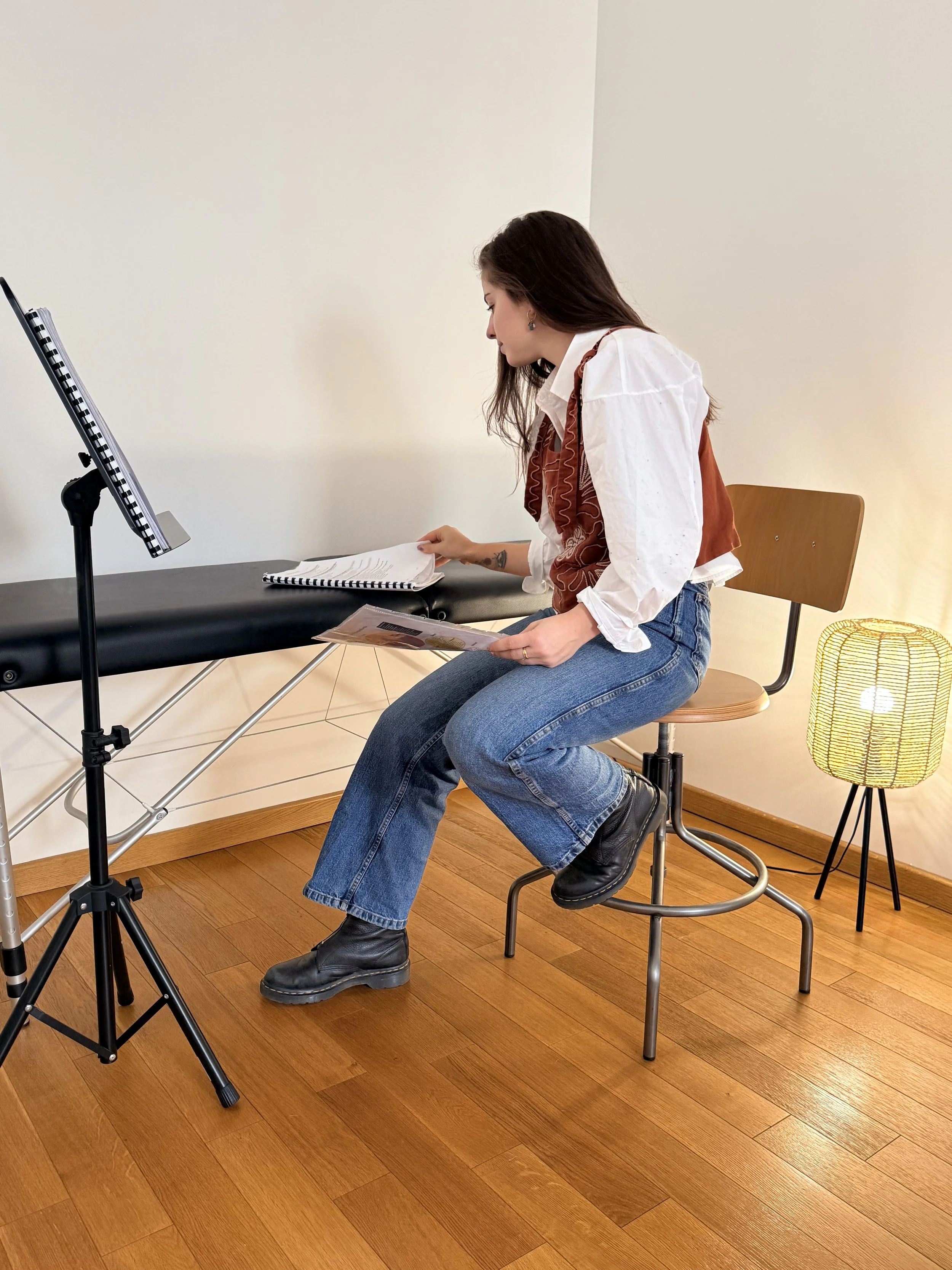 Une femme assise sur une chaise en studio, regardant des feuilles et des documents sur une table, avec un éclairage chaud, parquet en bois, et une lampe à côté.