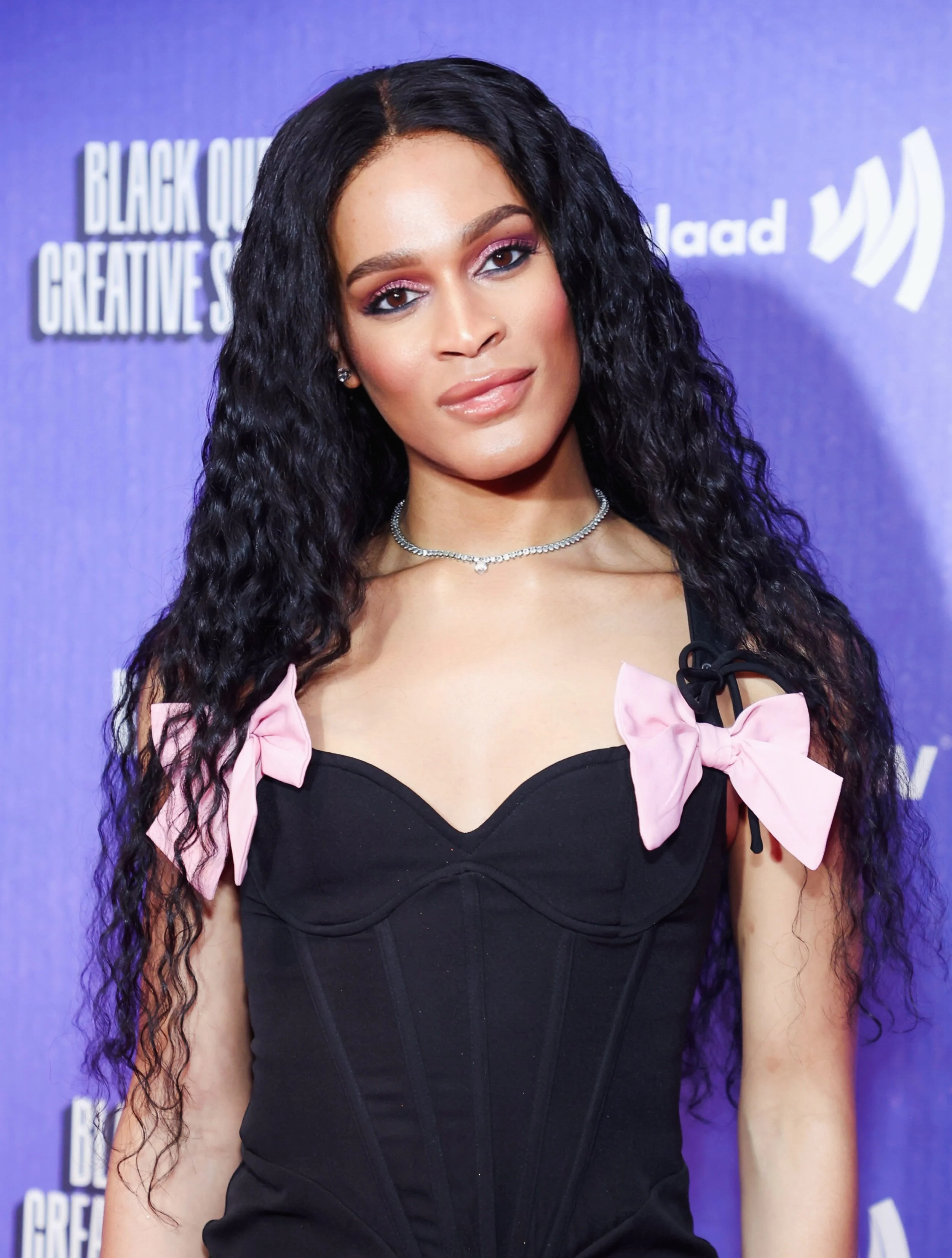 A woman with long, curly black hair and makeup, wearing a black dress with pink bows on the shoulders, a silver necklace, and earrings, at an event with a purple backdrop.