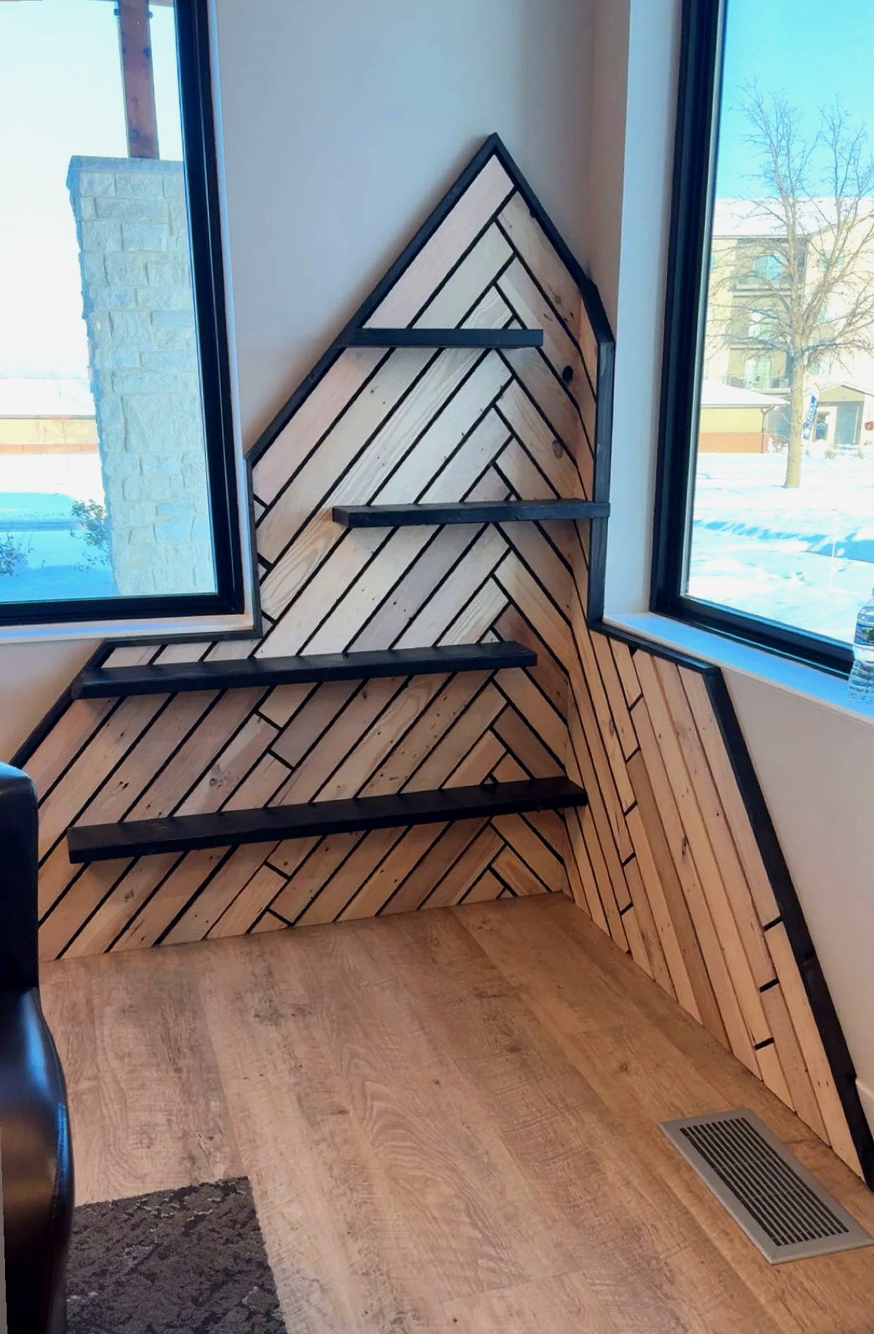 A wooden wall with diagonal planks arranged in a chevron pattern, bordered with black trim, featuring three black floating shelves, located in front of a large corner window showing a snowy outdoor scene.