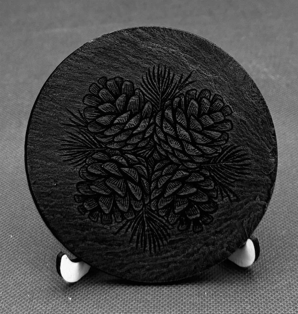 A round black board with an engraved pine cone design, displayed on a small stand against a textured gray background.