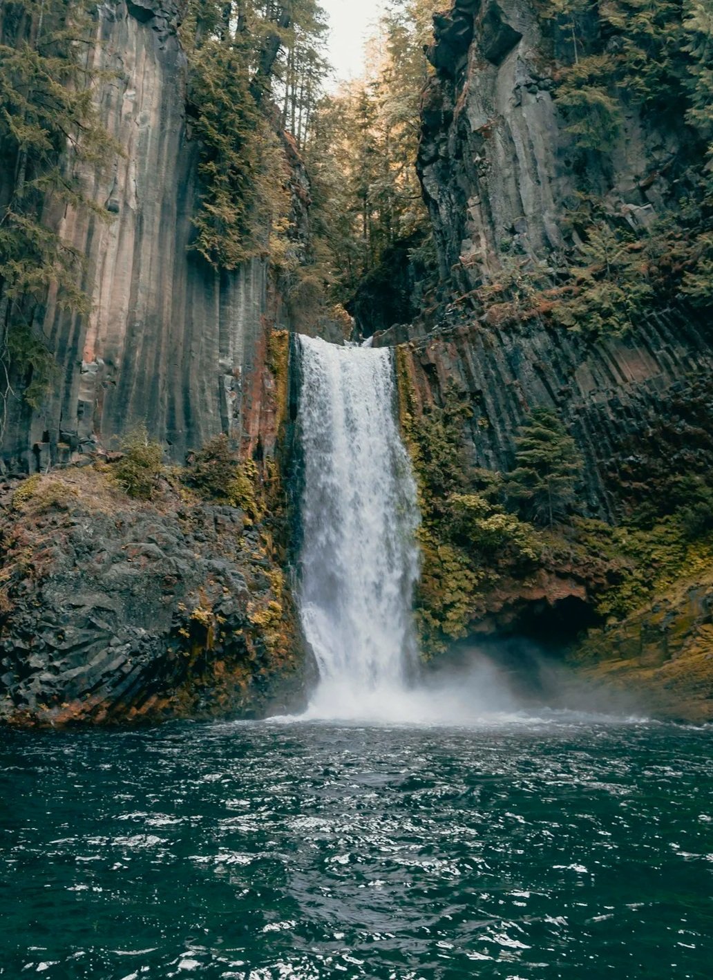 A tall waterfall cascading down dark, rugged cliffs surrounded by lush green trees, with water splashing into a pool below.